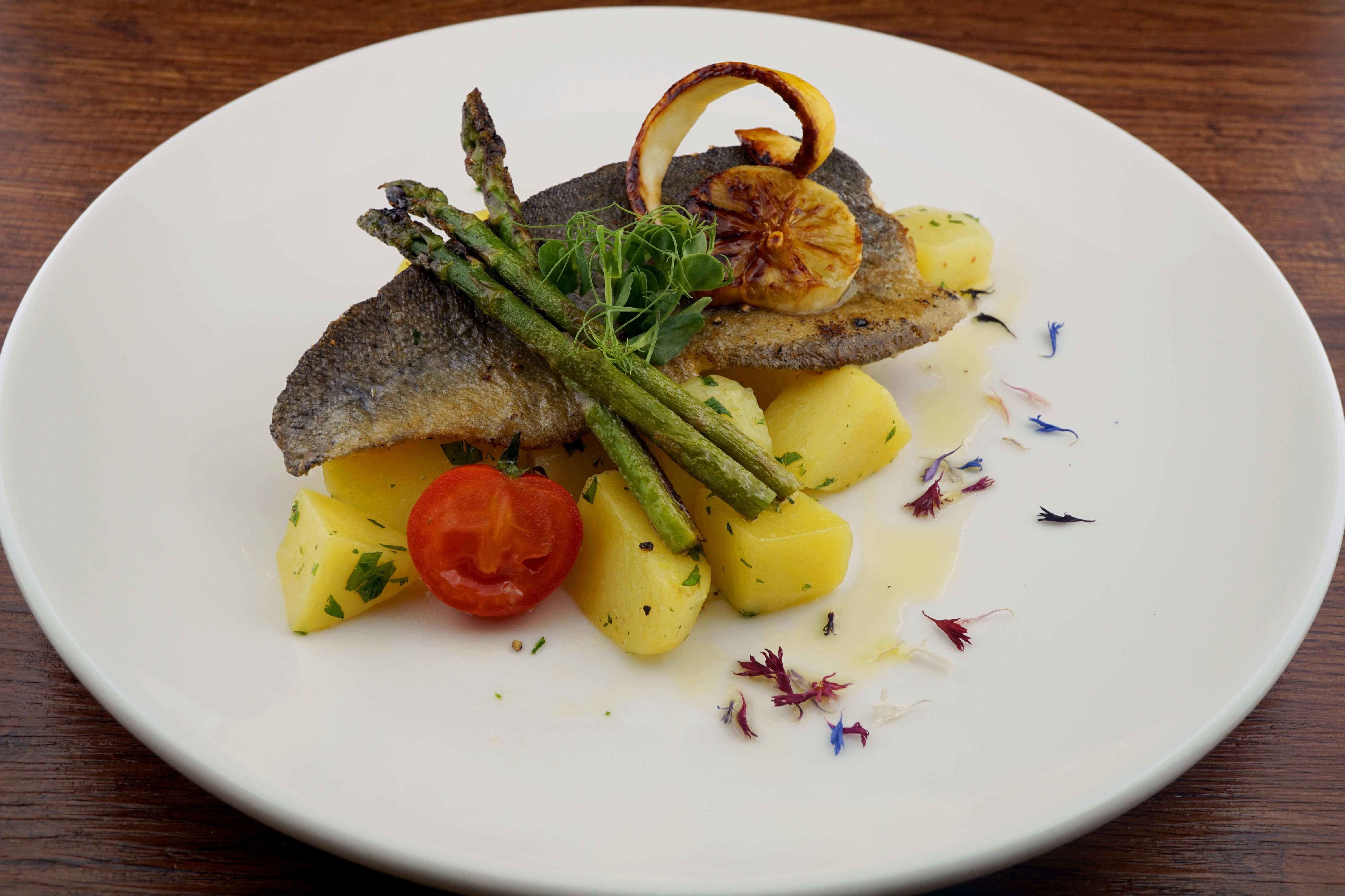 A delicious dish with fried fish, surrounded by fresh potatoes, asparagus, and a red tomato. The whole is decorated with herbs and edible flowers.