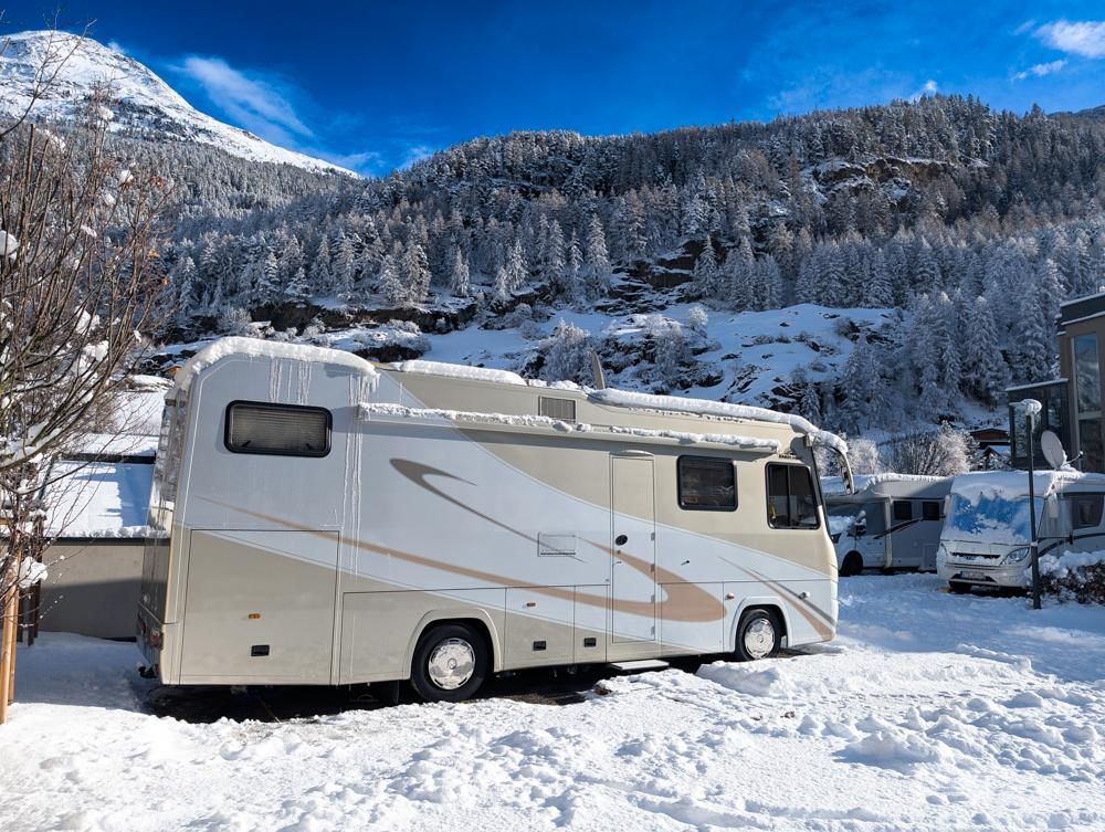 A motorhome is standing in a snowy landscape. In the background, snow-covered mountains and trees can be seen.