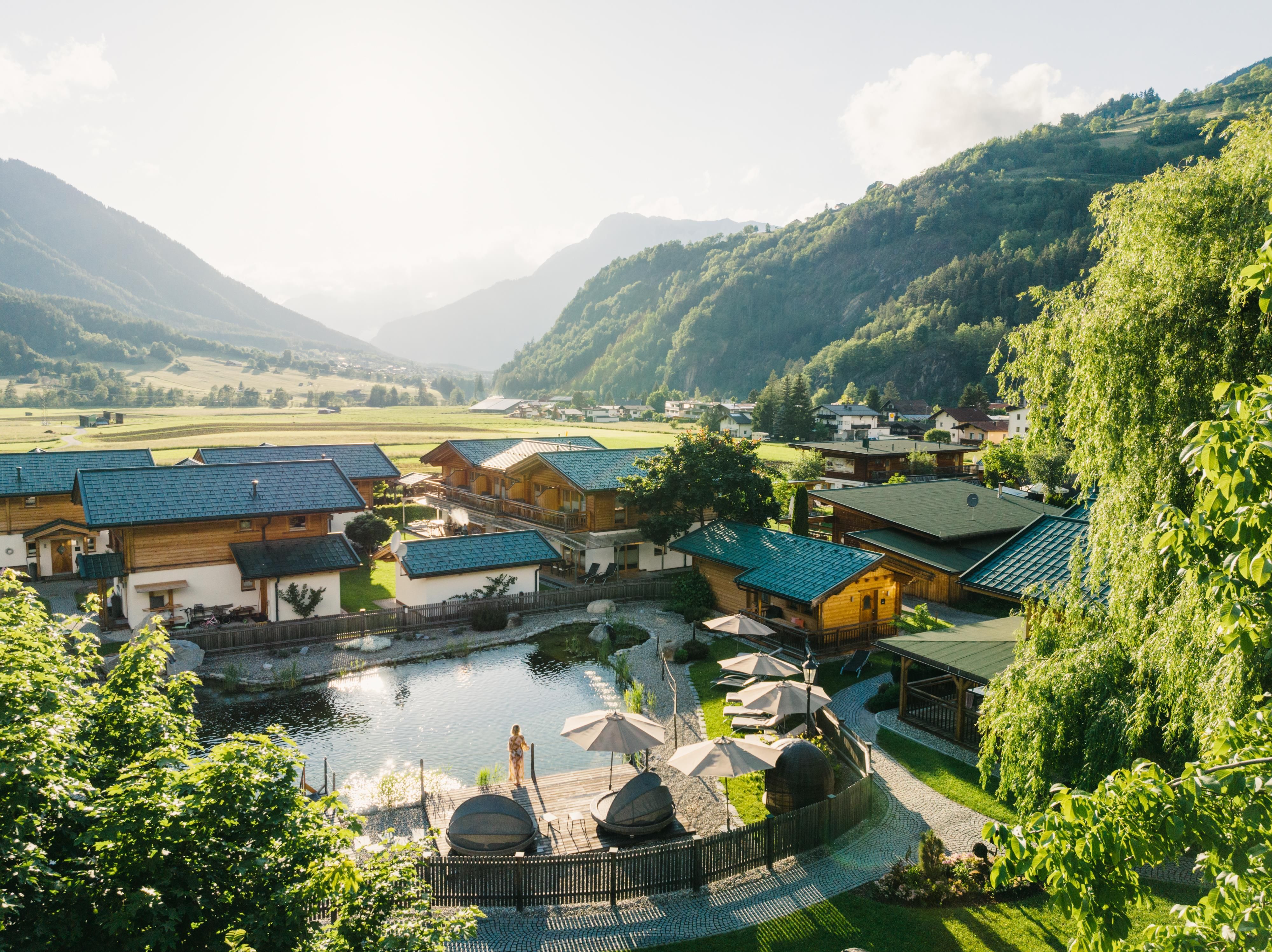 A picturesque mountain landscape with a swimming pond and cozy wooden cabins. The sun is shining and there is a calm, peaceful atmosphere.