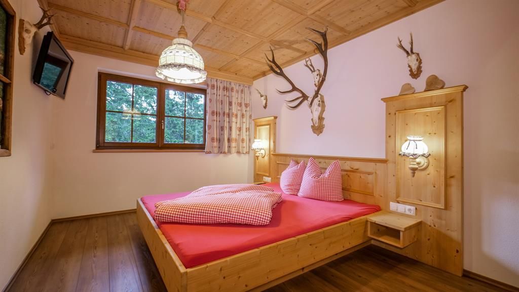 A cozy bedroom with a large red bed and wooden paneling. Antlers hang on the walls, and there is a window with a view outside.