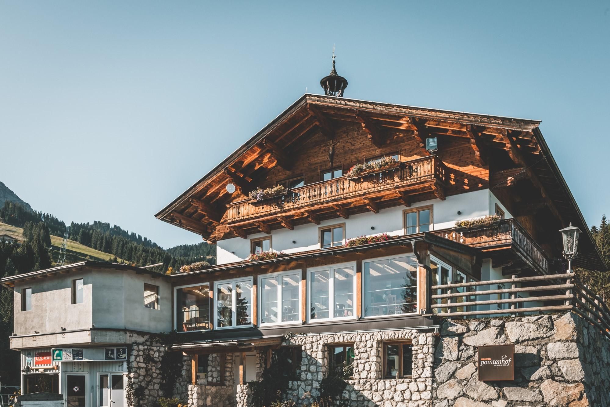 A traditional alpine house with wooden cladding and a balcony. In the background, green mountains can be seen.