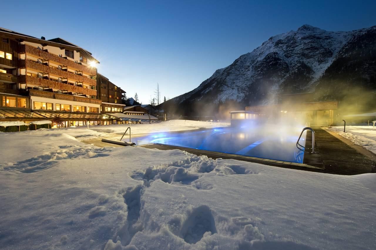 A heated outdoor pool surrounded by snow and mountains. In the background, there's a cozy building at night.