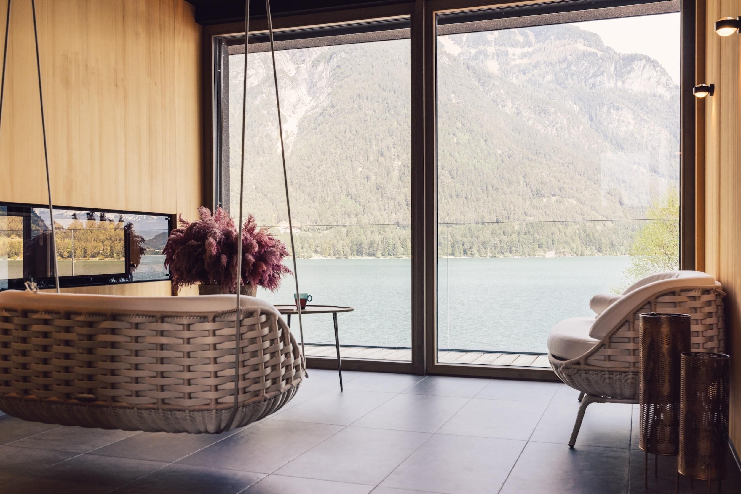 A modern living room with a swinging chair and large windows that offer a view of a lake and mountains. The decor is minimalist and elegant.