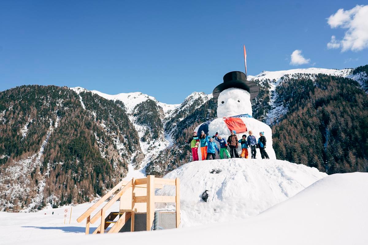 A group of children is standing next to a large snowman in a winter landscape. In the background, snow-covered mountains and a blue sky can be seen.
