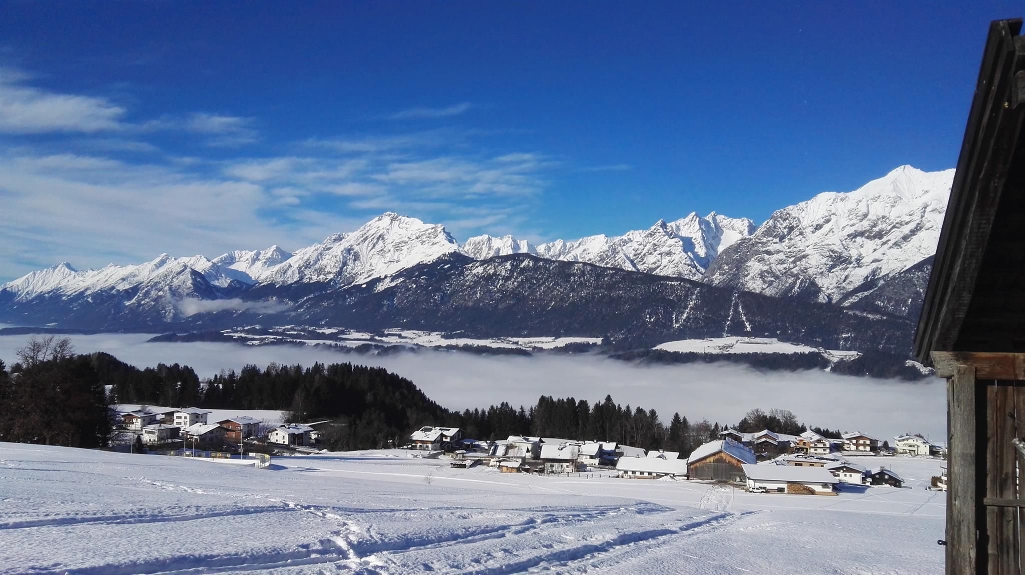 A snowy mountain landscape with tall peaks and a clear blue sky. In the foreground, there are snow-covered huts and a valley with fog.