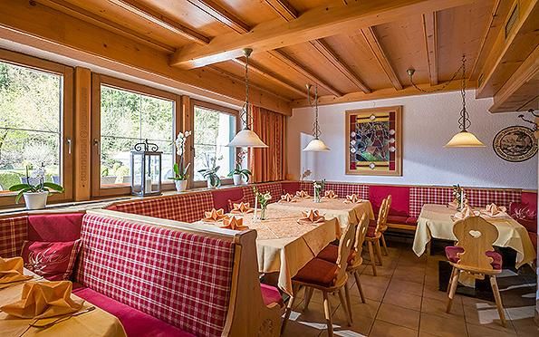 A cozy restaurant with a wooden beam ceiling and red checkered chairs. The tables are decorated with yellow tablecloths and the windows offer a view of greenery.