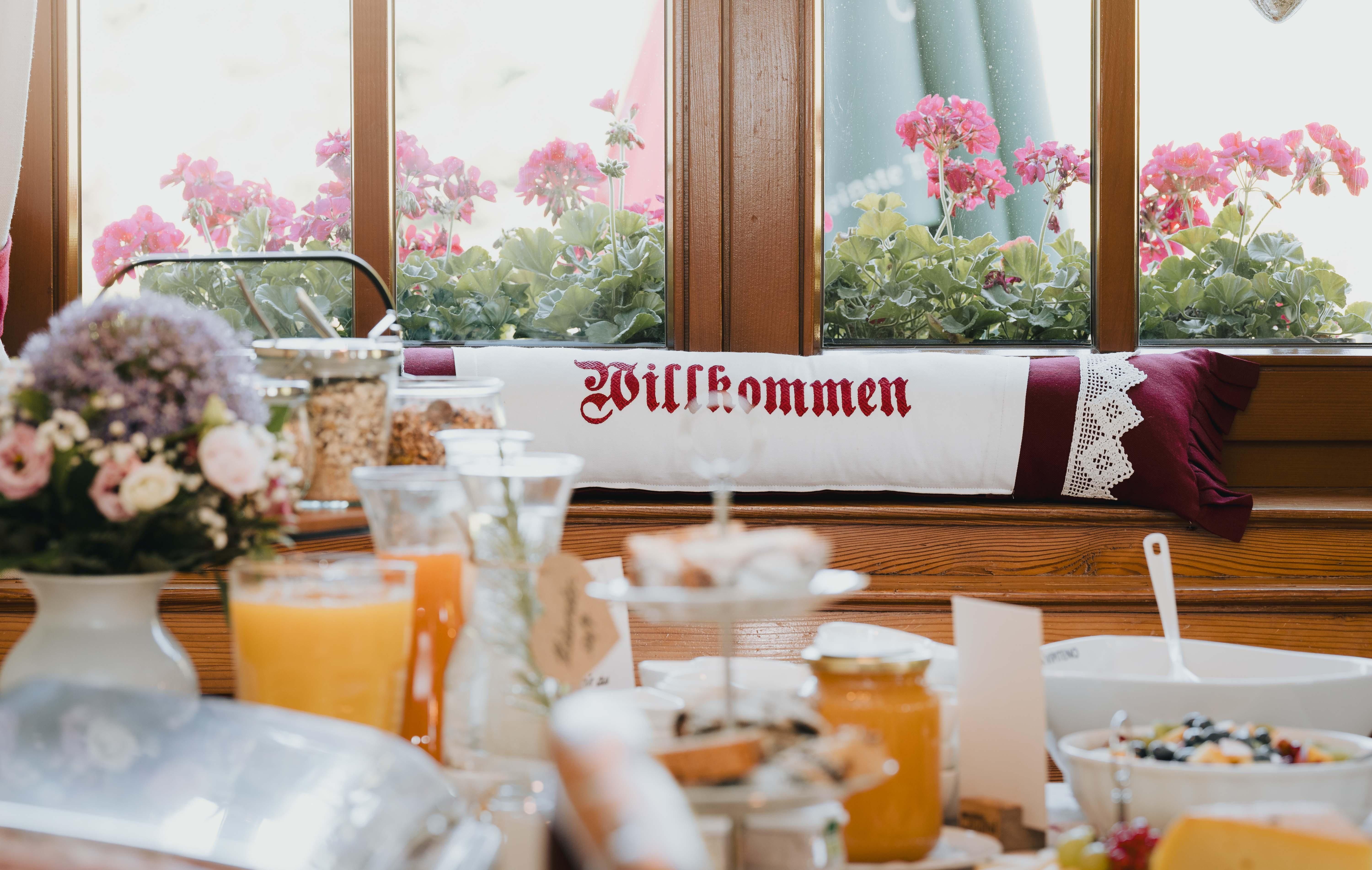 A nicely set table with breakfast offerings and a welcome sign. In the background, blooming plants can be seen.