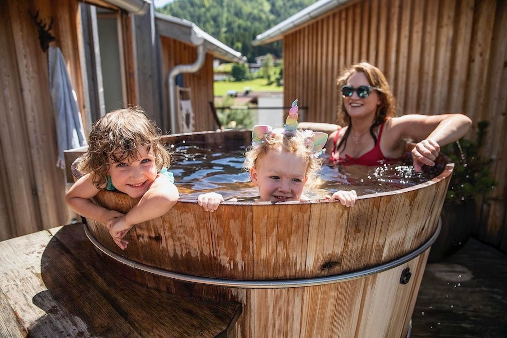 A mother with her two children relaxes in a wooden bath. They are enjoying a sunny day outdoors.