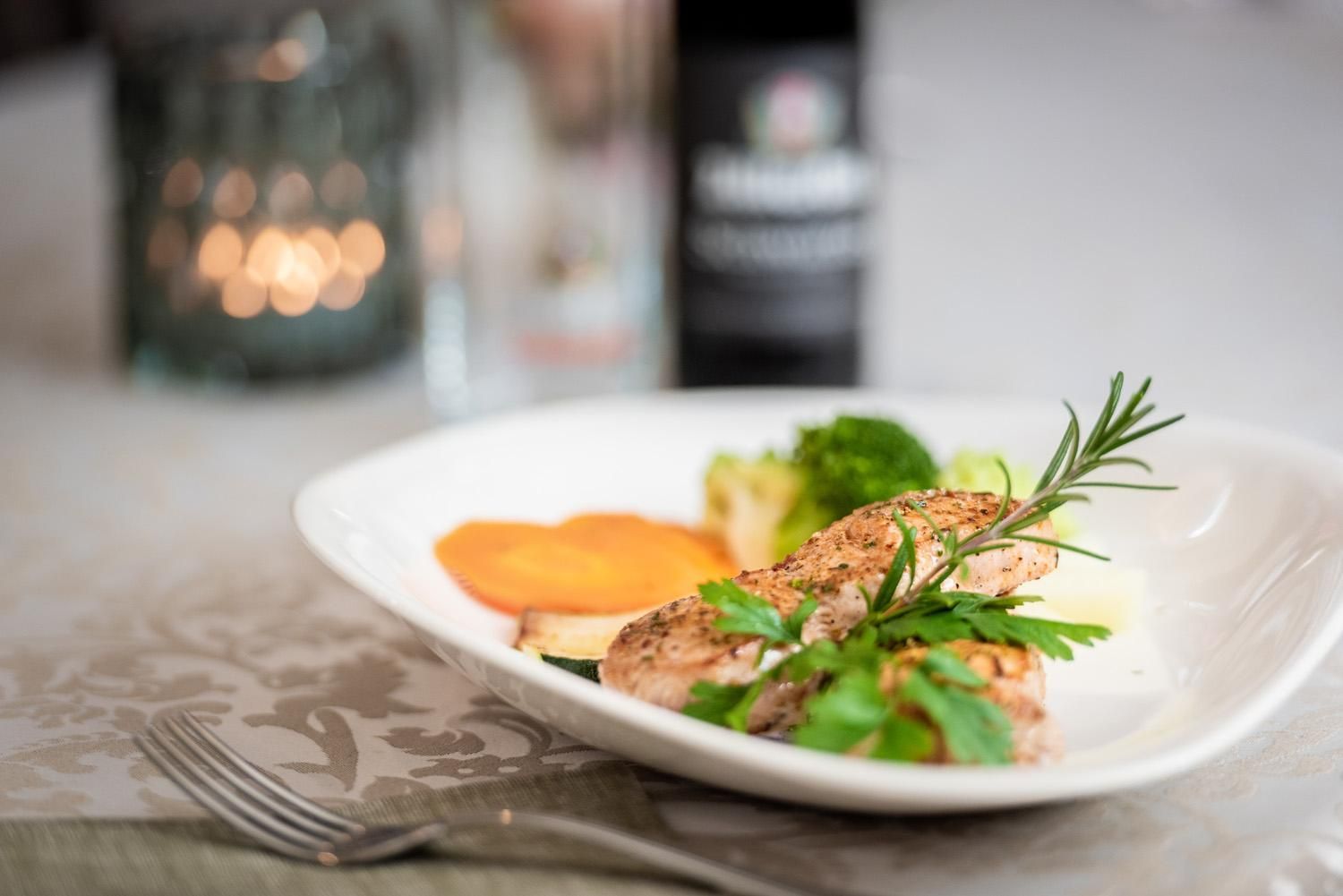 An attractively arranged plate with grilled fish, colorful vegetables, and fresh herbs. In the background, a candle and a bottle of wine are visible.