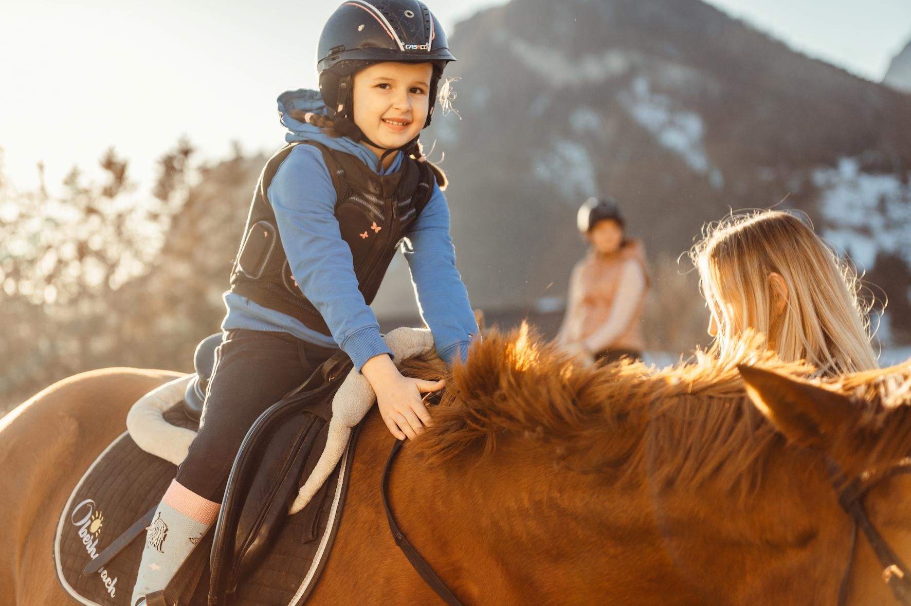 A smiling child is riding on a brown horse and stroking its mane. In the background, trees and mountains can be seen, creating a peaceful atmosphere.