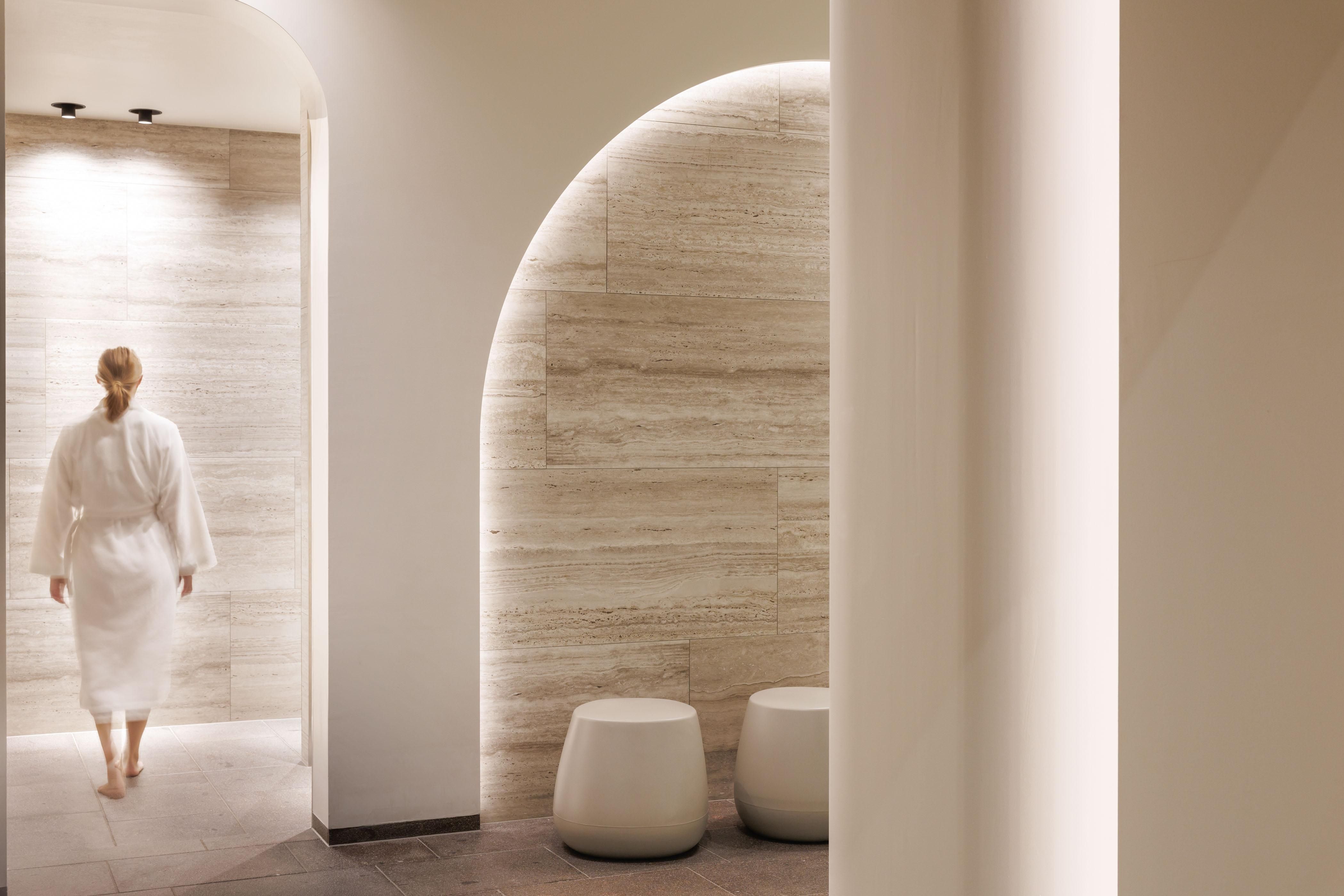 A person in a bathrobe walks into a modern spa area. The walls are bright and softly lit, with elegant seating.