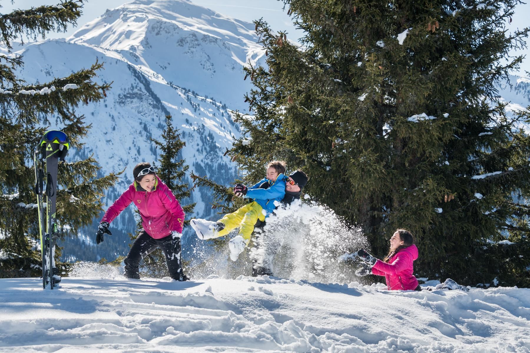 A happy group of children is playing in the snow. In the background, there are snow-covered mountains and green trees.
