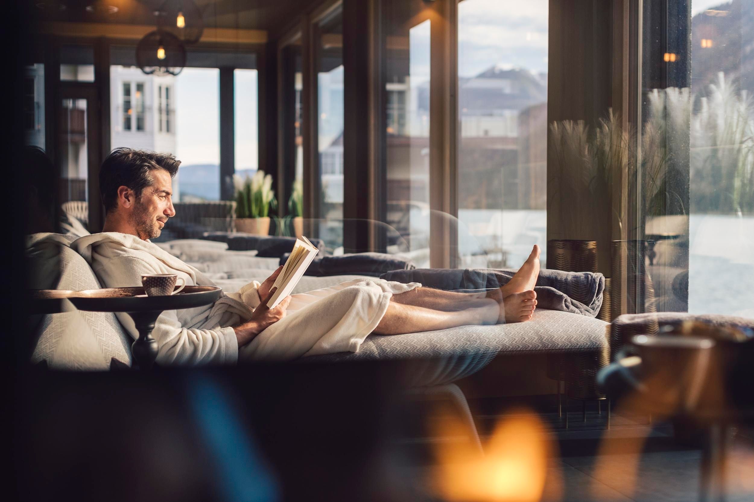 A man is sitting relaxed in a bathrobe on a couch and reading a book. The windows offer a view of the landscape outside.