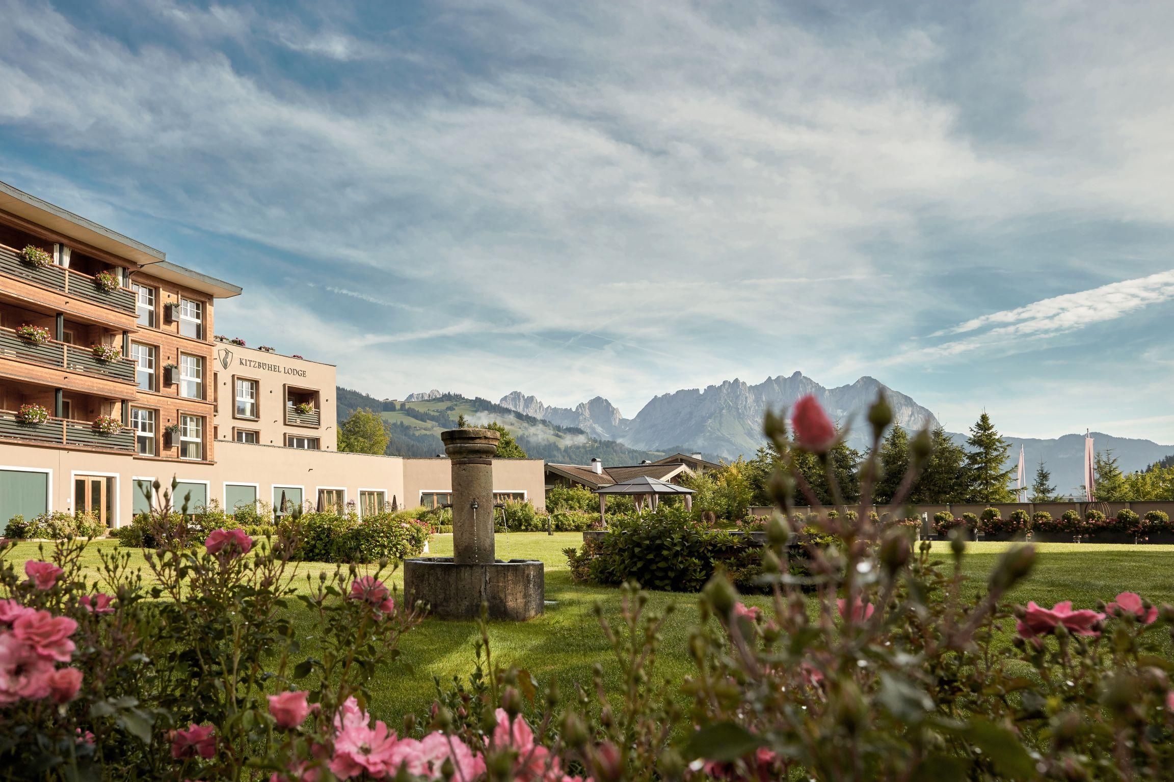A beautiful hotel building with a well-maintained garden full of roses. In the background, majestic mountains and a clear sky can be seen.