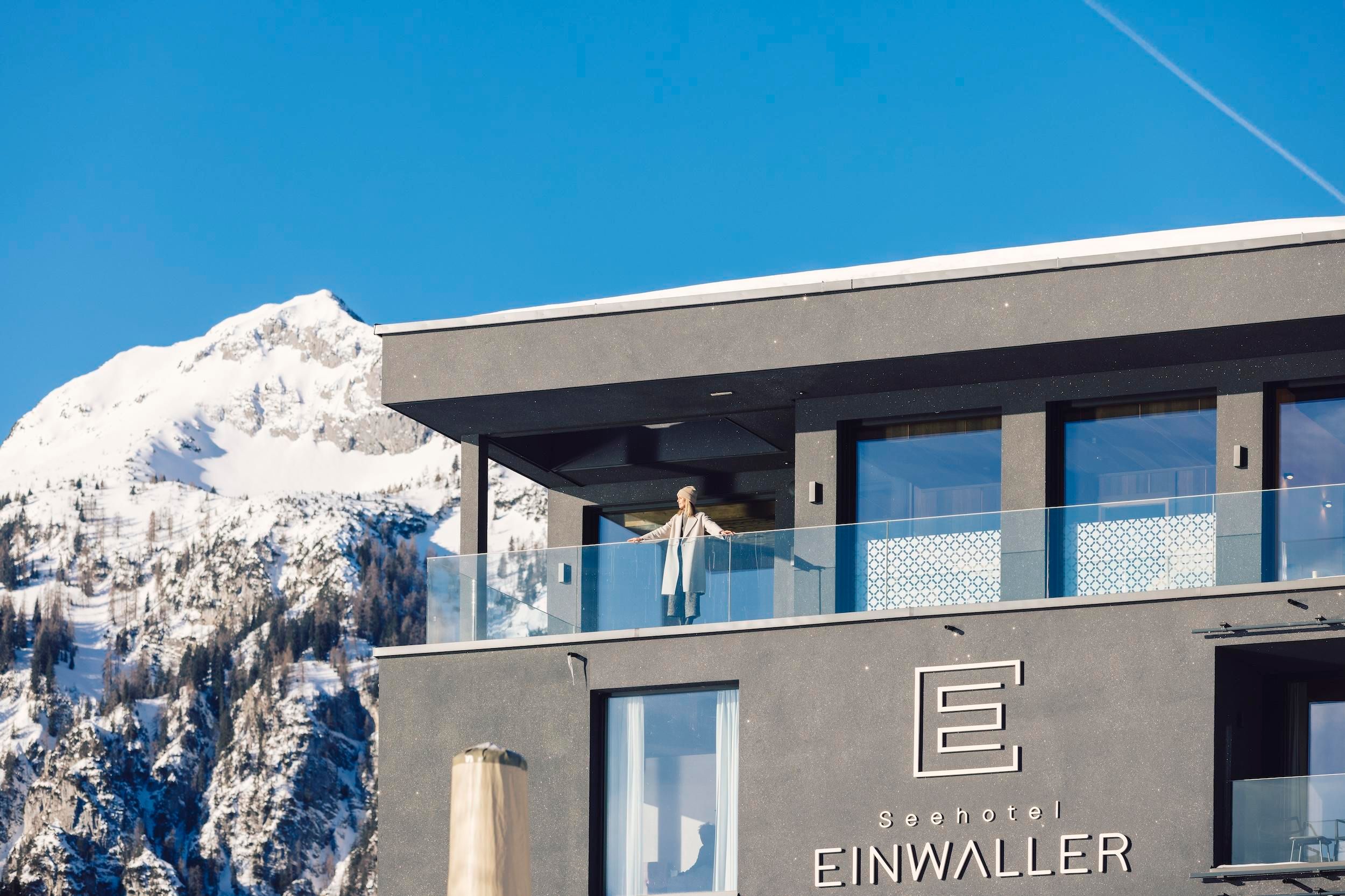 A modern hotel with a balcony and a person enjoying the view. In the background, snow-covered mountains and a clear blue sky can be seen.