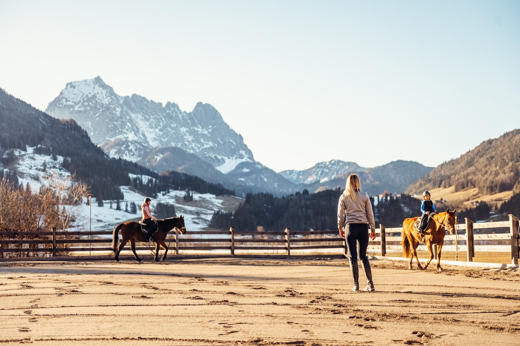 A rider on a brown horse and another person on a white horse are riding in a picturesque landscape. In the background, snow-capped mountains and a blue sky can be seen.
