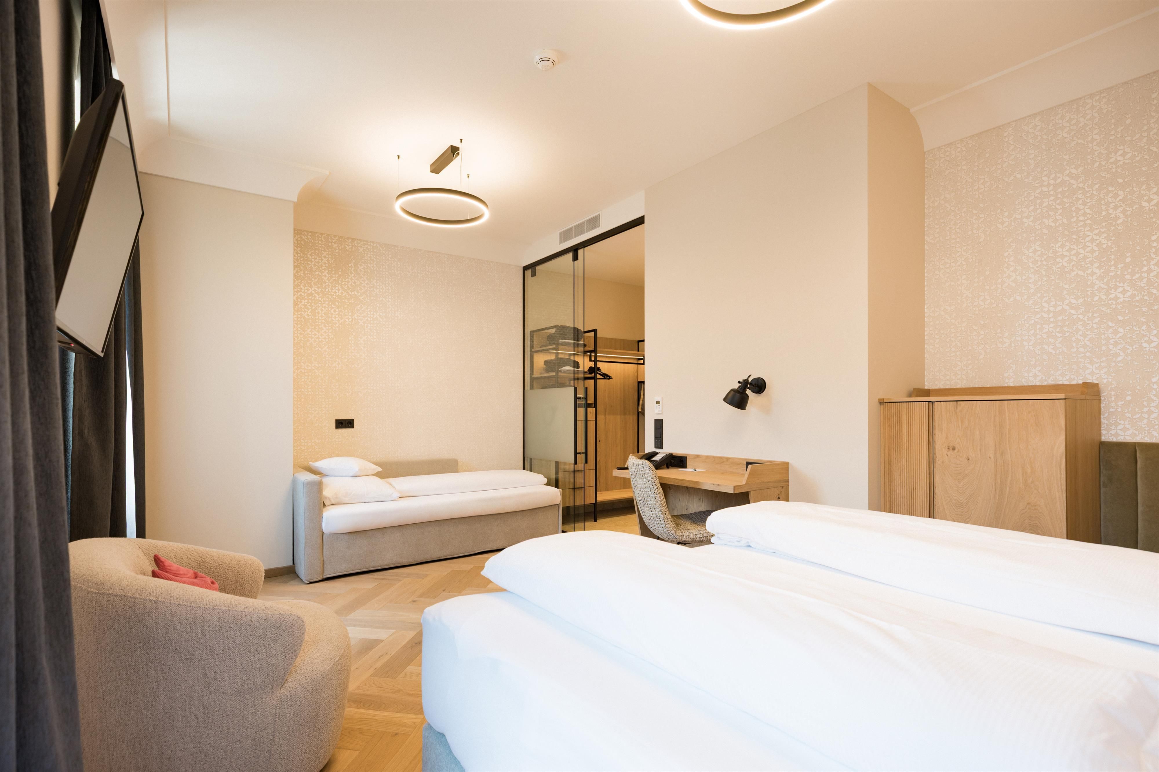 A modern hotel room with two beds and a comfortable chair. The walls are painted in neutral tones, and the room feels bright and inviting.