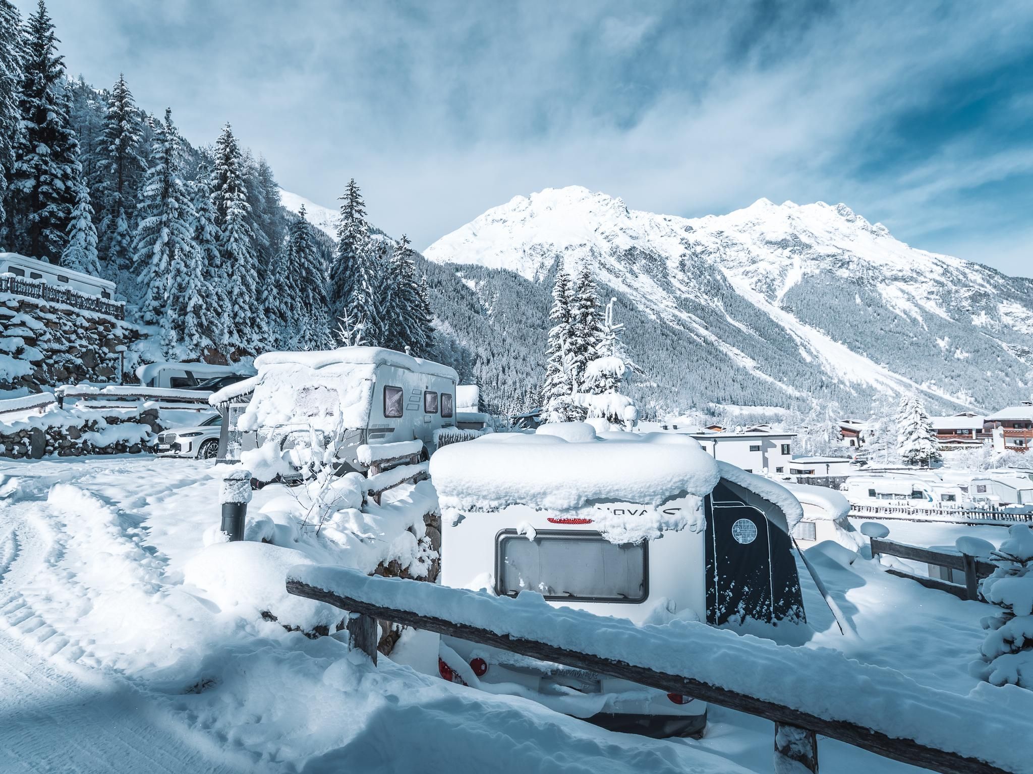 A snow-covered landscape with RVs, surrounded by tall trees and mountains. The sky is clear and blue.