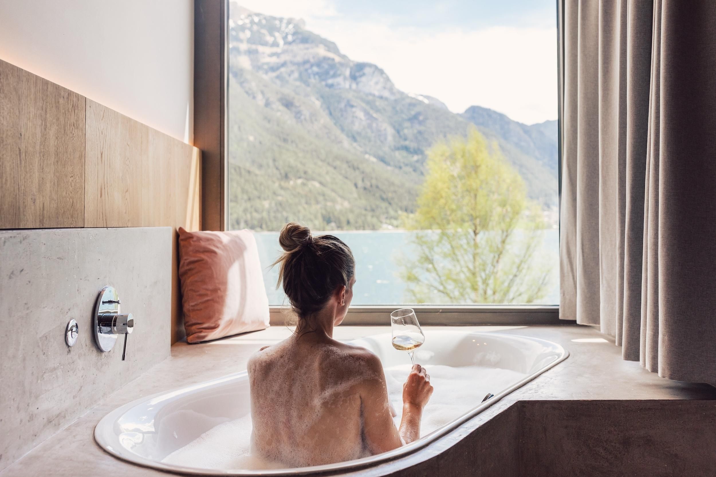 A person relaxes in a bathtub with a view of the mountains and the lake. They hold a glass of wine and enjoy the tranquil atmosphere.