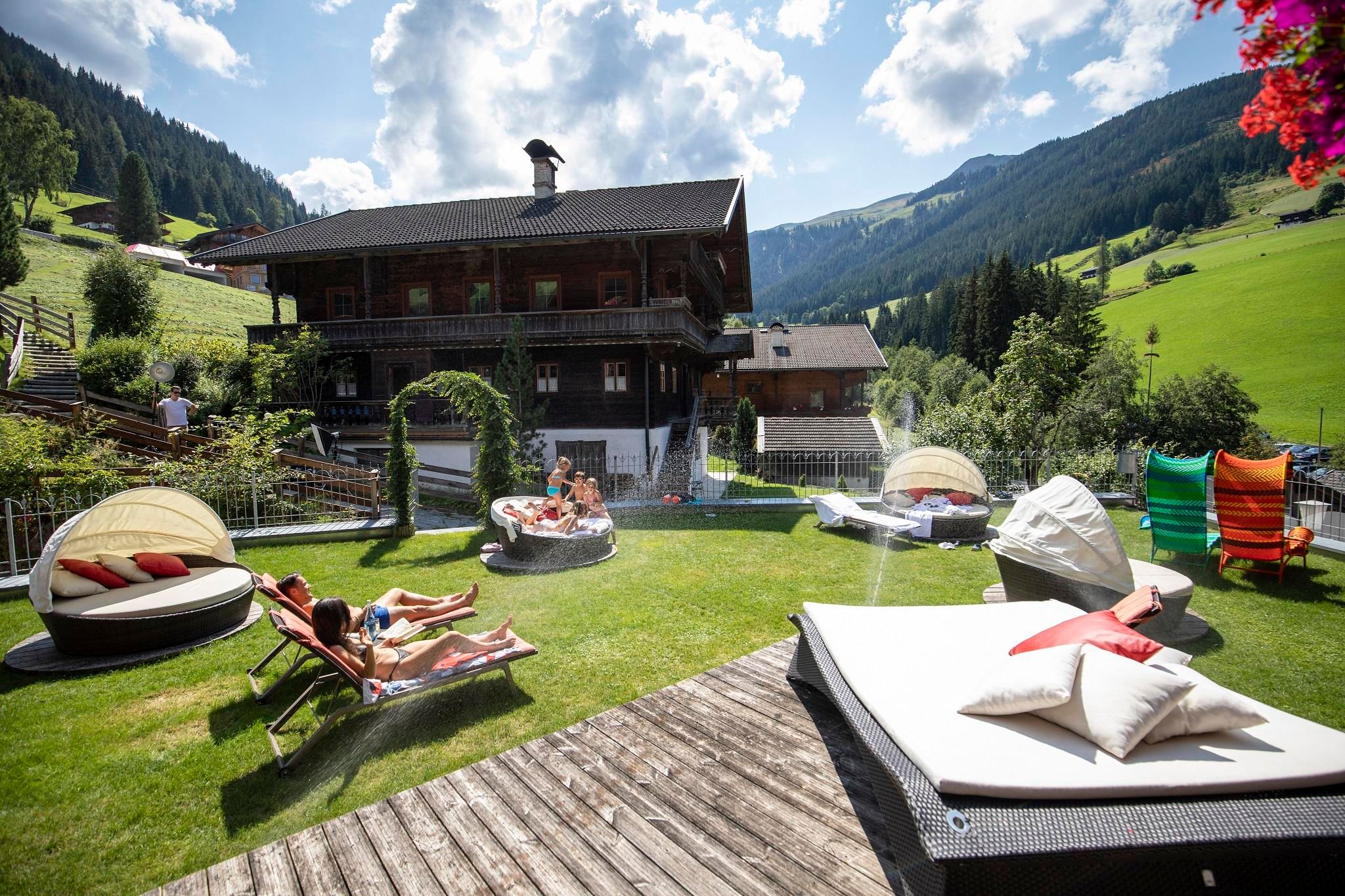 A picturesque mountain house surrounded by green meadows. In the foreground, people are relaxing on loungers under a bright blue sky.