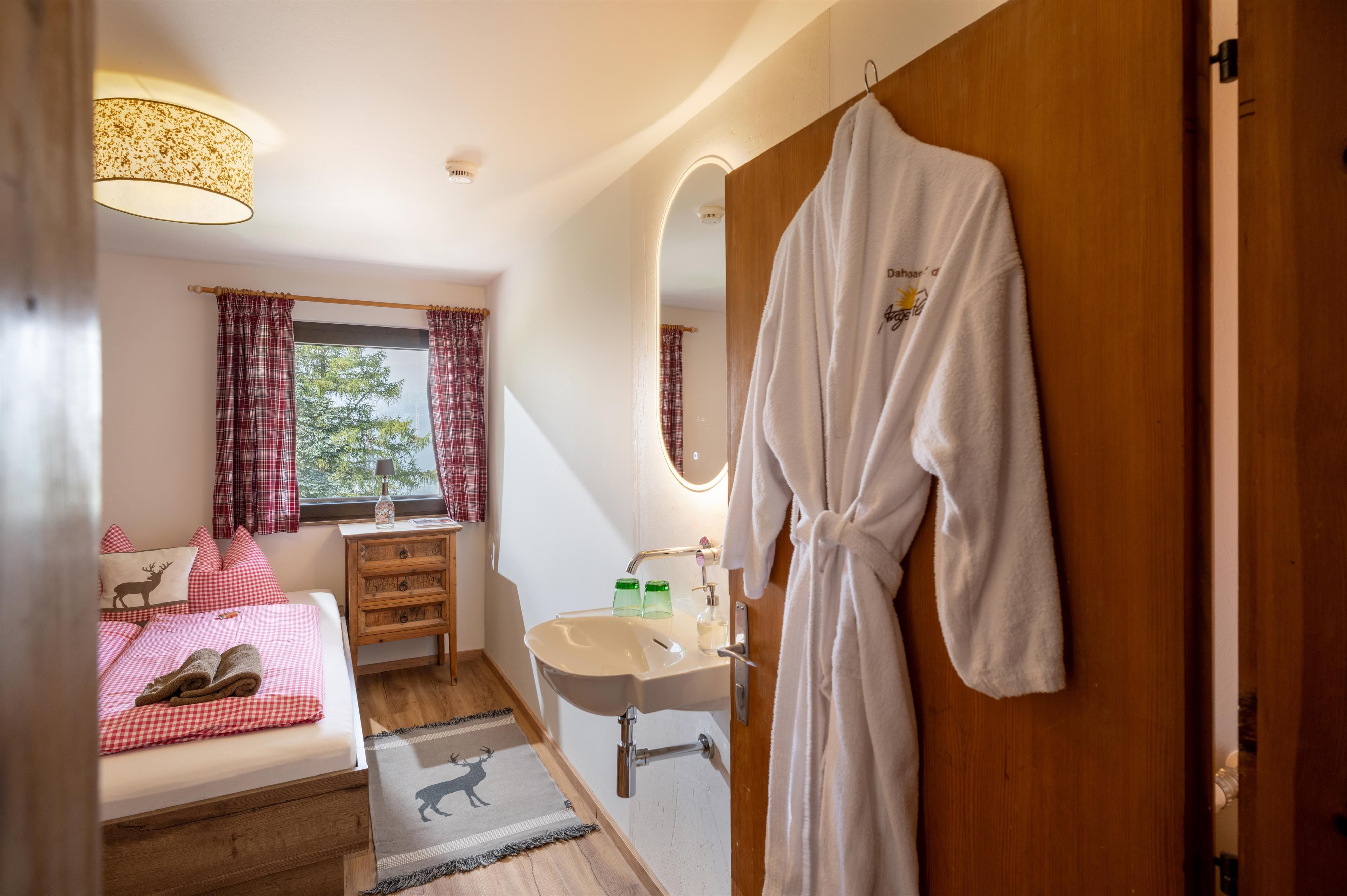 A cozy guest room with a double bed and a window that lets in a lot of light. A bathrobe hangs on the door, and there is a small washbasin in the room.