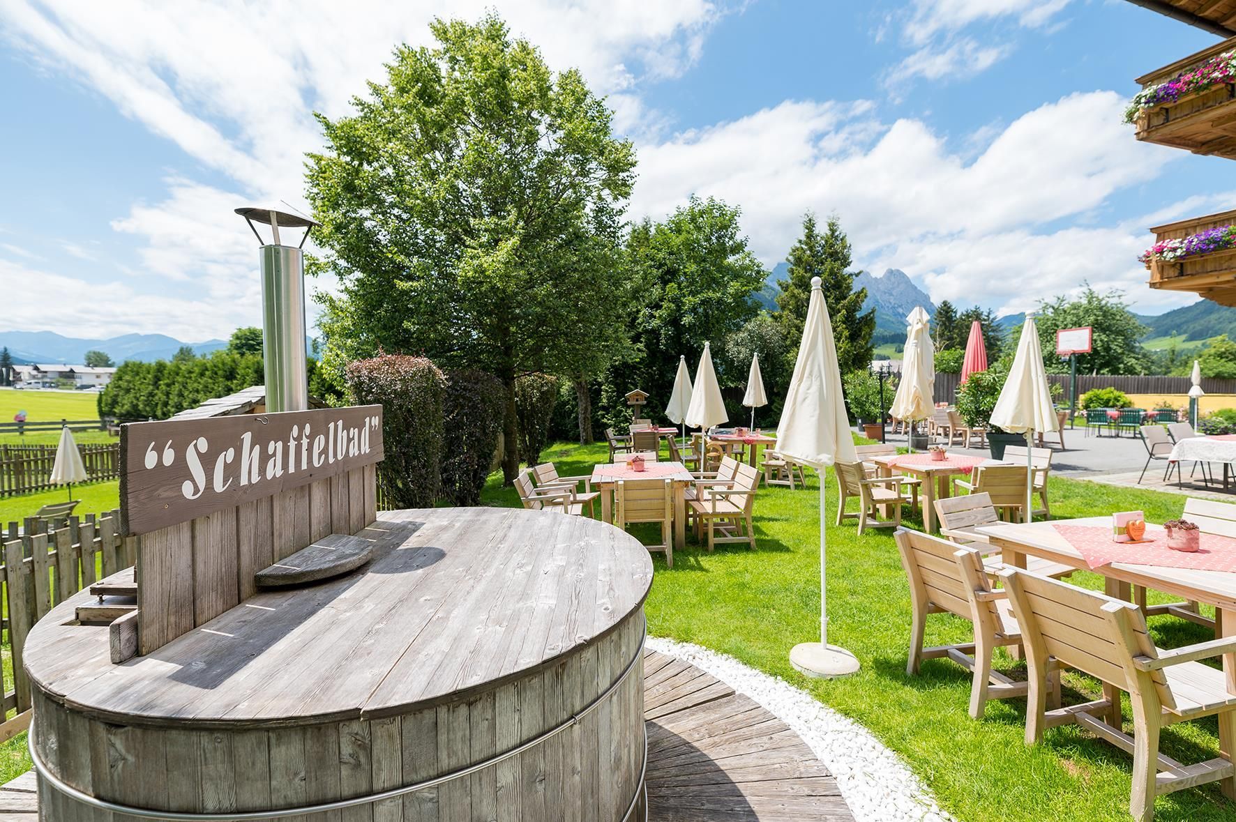 An inviting garden with wooden tables and sun umbrellas. In the background, there are trees and a beautiful landscape with mountains.