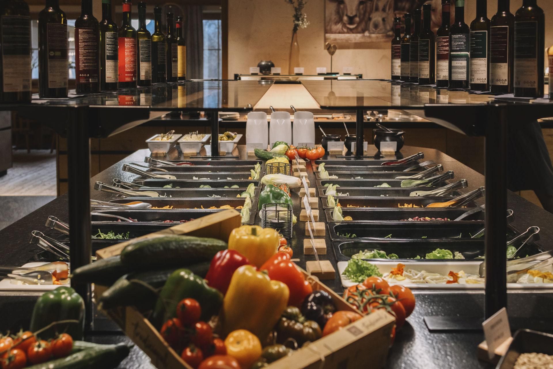 A colorful selection of fresh vegetables and salad in a buffet area. In the background, bottles of various oils and vinegars are displayed.