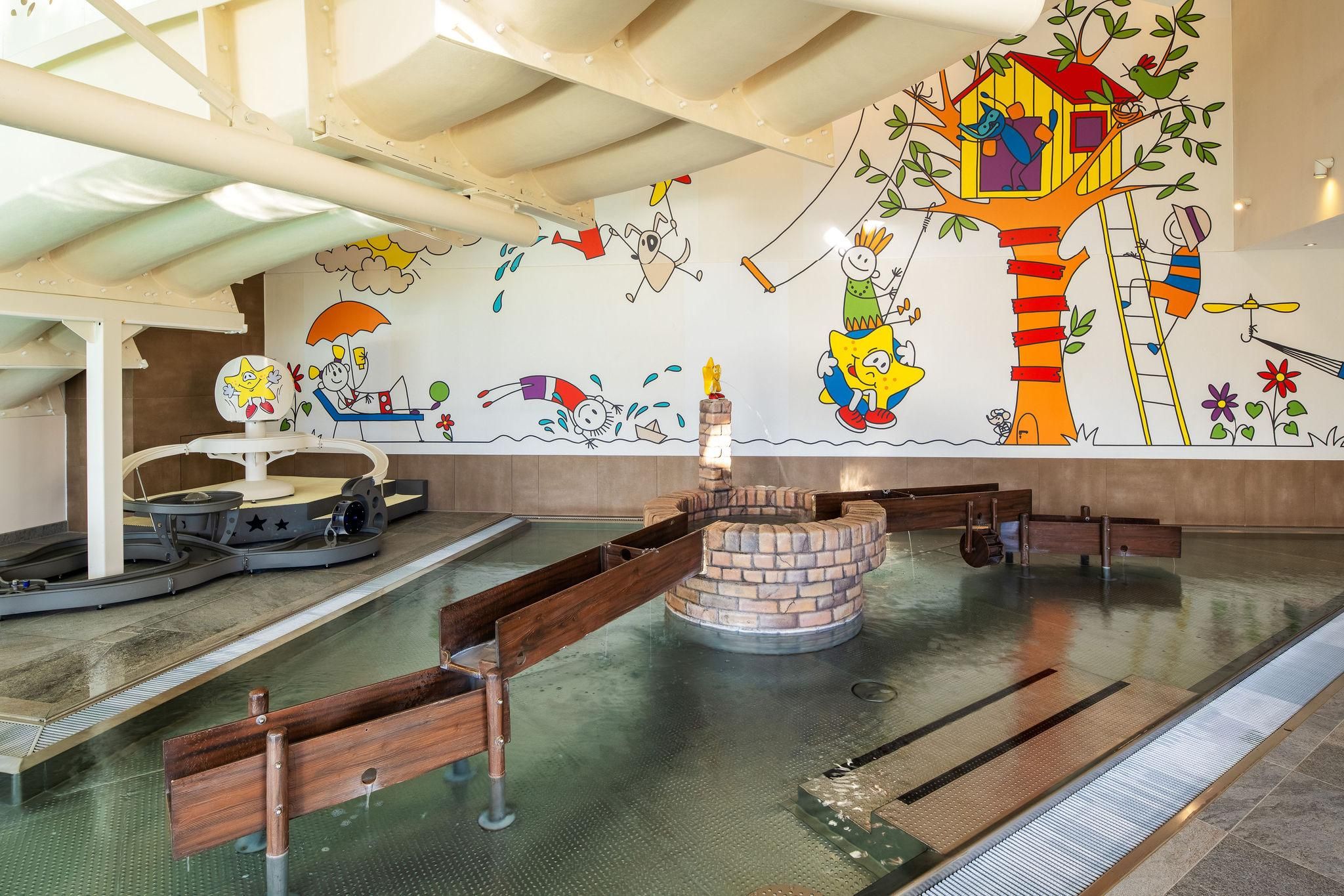 Children's pool with a water play area. On the wall are colorful, kid-friendly drawings of a treehouse and cheerful characters.