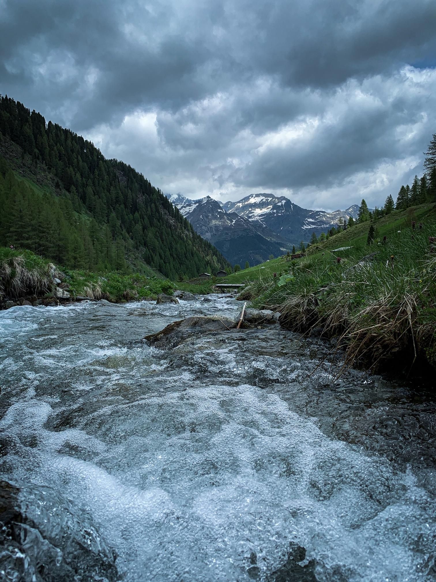 A clear river flows through a green mountain landscape. In the background, snow-covered mountains and a cloudy sky can be seen.