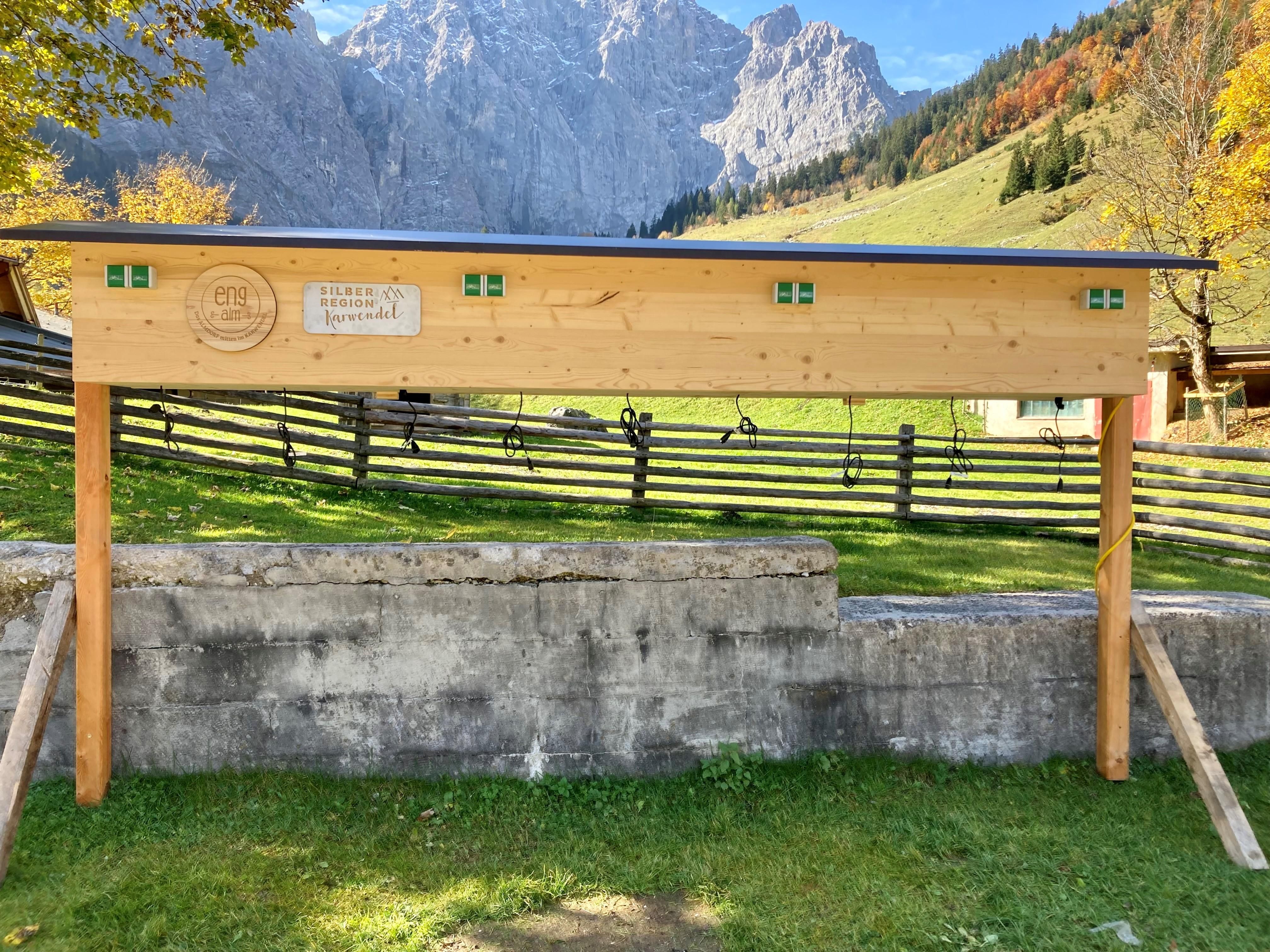 A wooden information board stands in front of a picturesque mountain landscape. In the background, there are mountains and a green meadow.