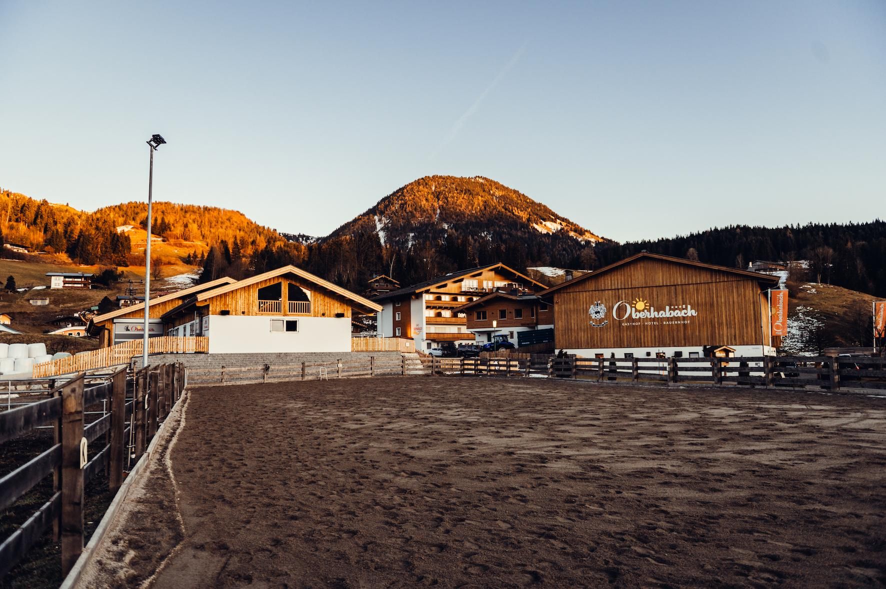 A landscape with an equestrian arena and traditional wooden houses. In the background, gentle mountains and a golden evening atmosphere can be seen.