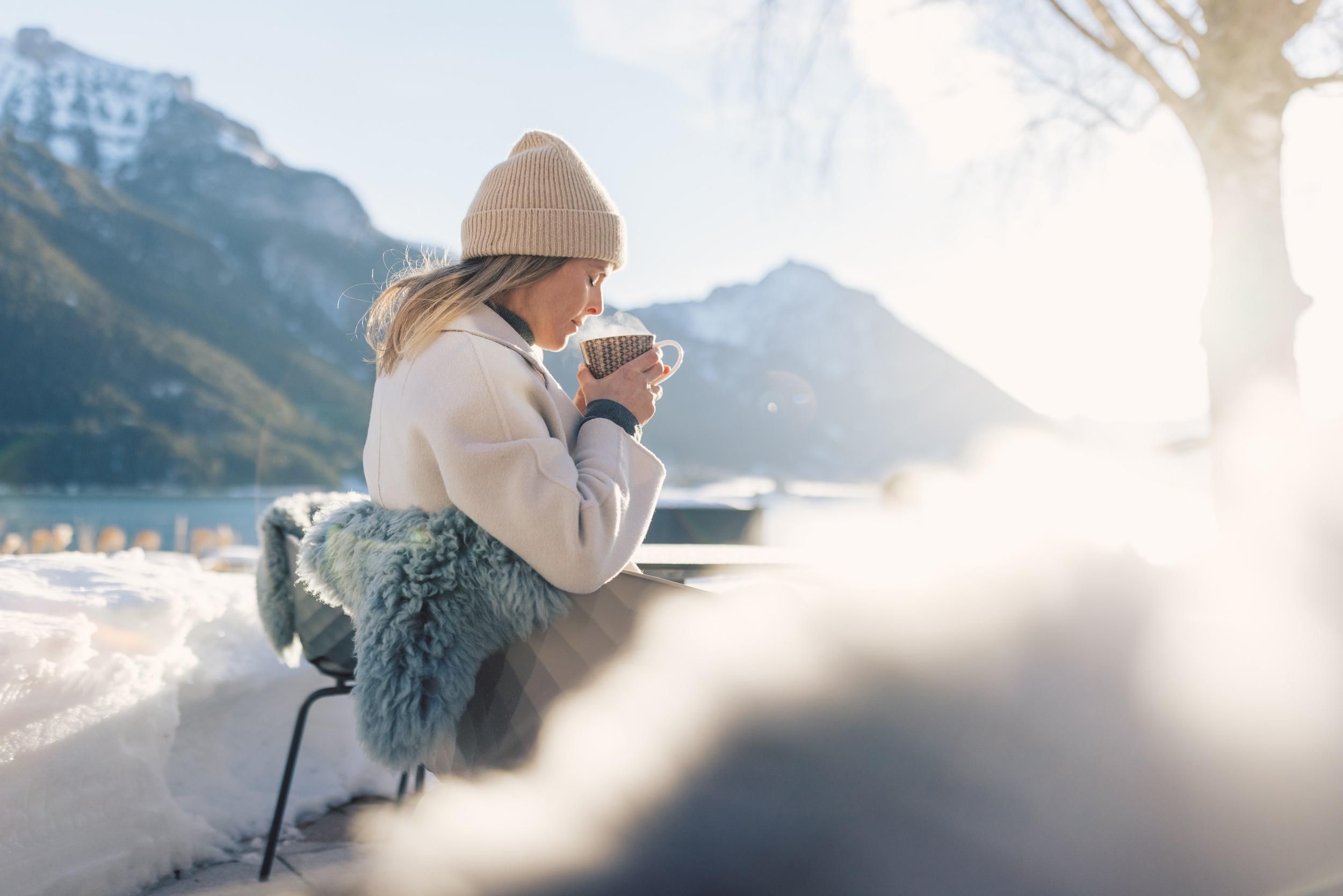 A woman is sitting outside and enjoying a hot drink, surrounded by snow and mountains. The sun is shining, creating a cozy atmosphere.