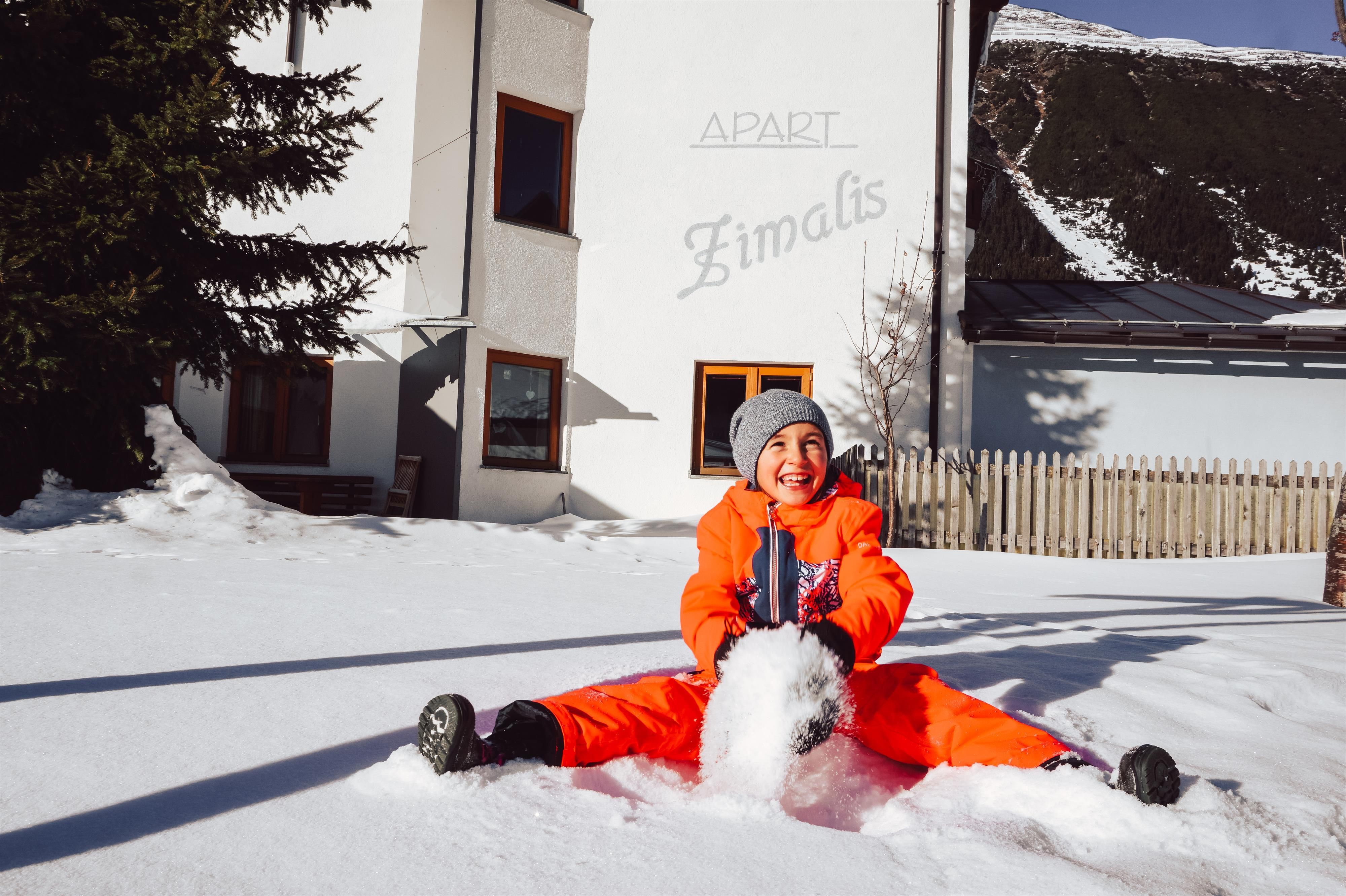 A happy child in orange winter clothing sits in the snow and builds a snowman. In the background, a holiday house and snow-covered trees can be seen.