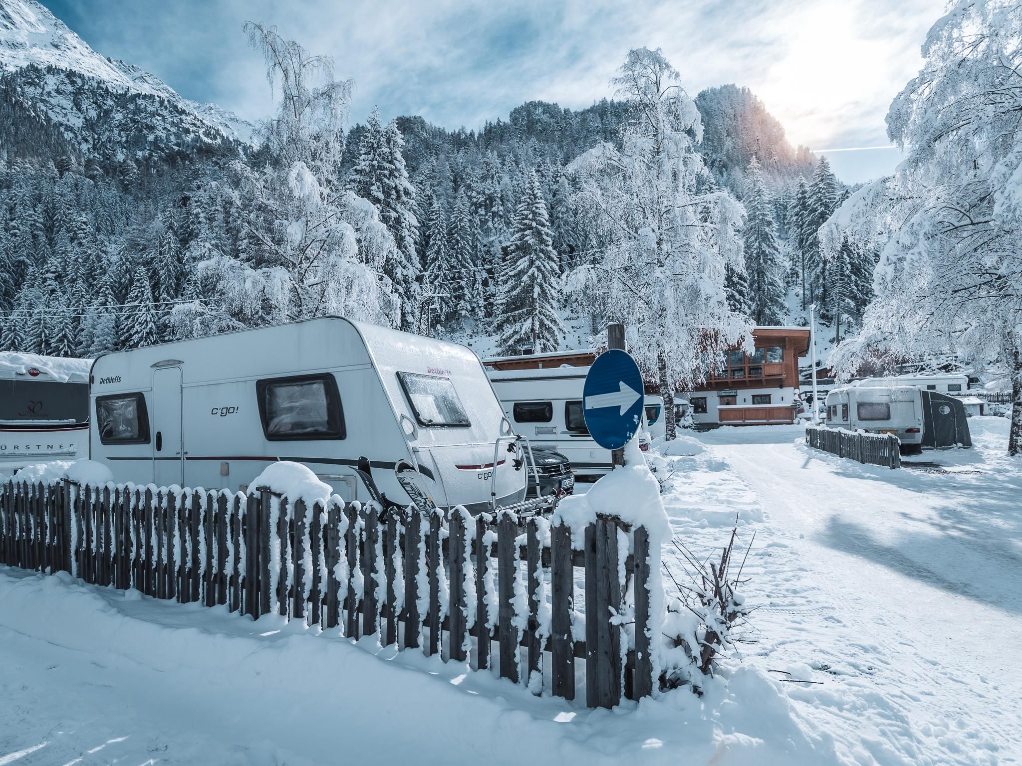 A snowy campsite with motorhomes and a wooden fence. In the background, snow-covered trees and mountains are visible.
