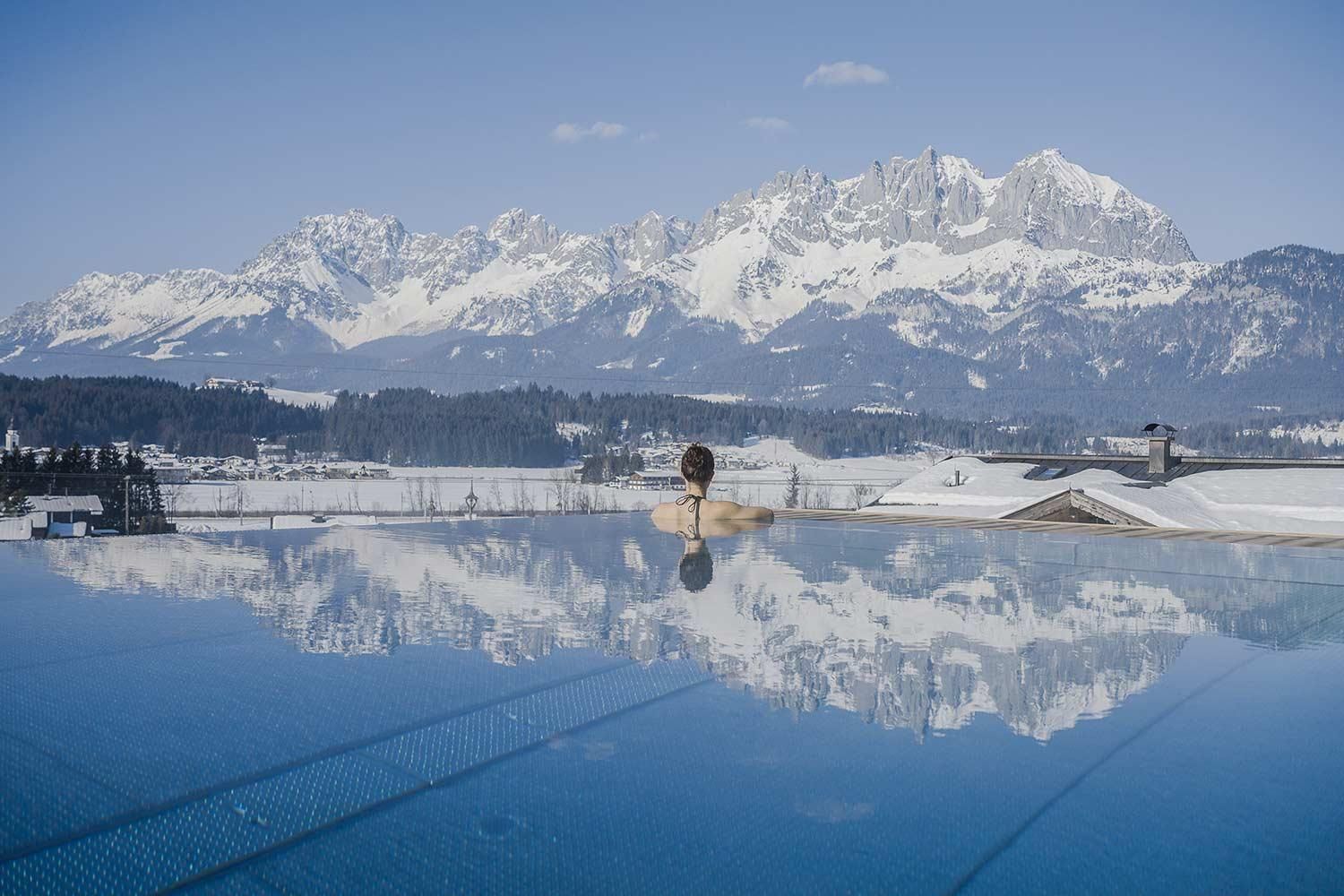 A breathtaking view of snow-covered mountains with a tranquil swimming pool in the foreground. The sky is clear and radiant blue.