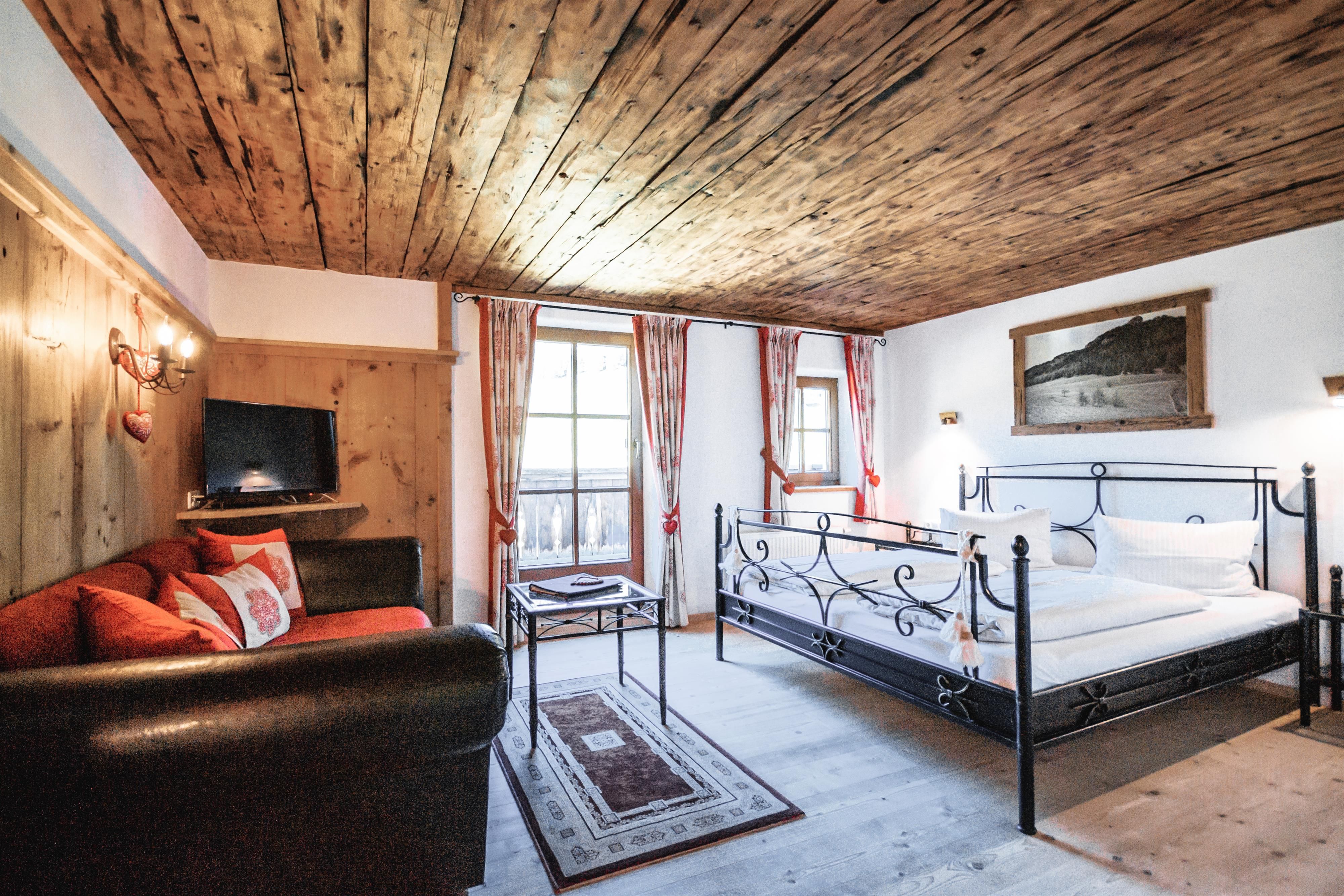 A cozy room with wooden walls and a double bed. There is a seating area with a black sofa and large windows that let in plenty of light.