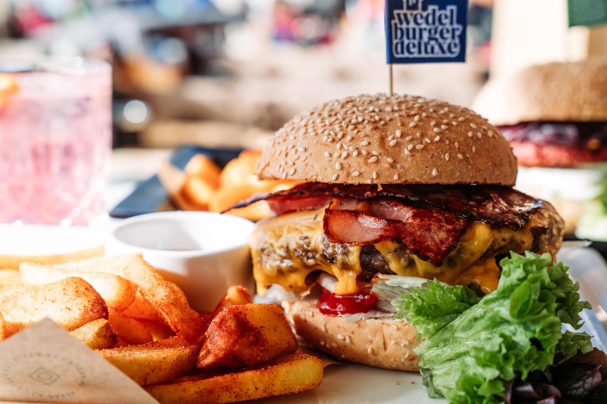 A juicy burger with a sesame bun, cheese, and bacon, garnished with a small flag toothpick. Served with crispy fries and a fresh salad.