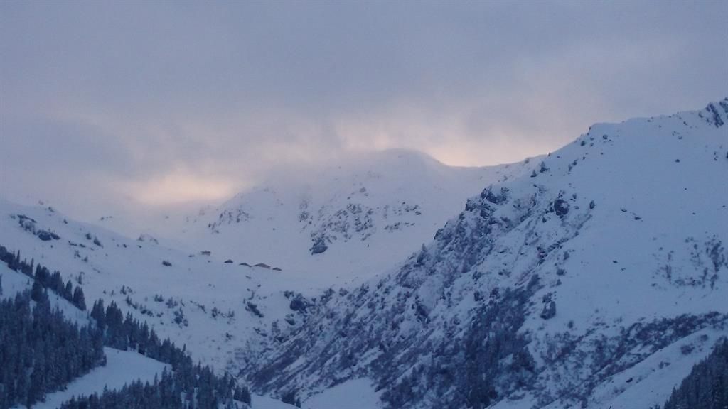 A snowy mountain landscape under a cloudy sky. Gentle hills and trees are visible in the foreground.