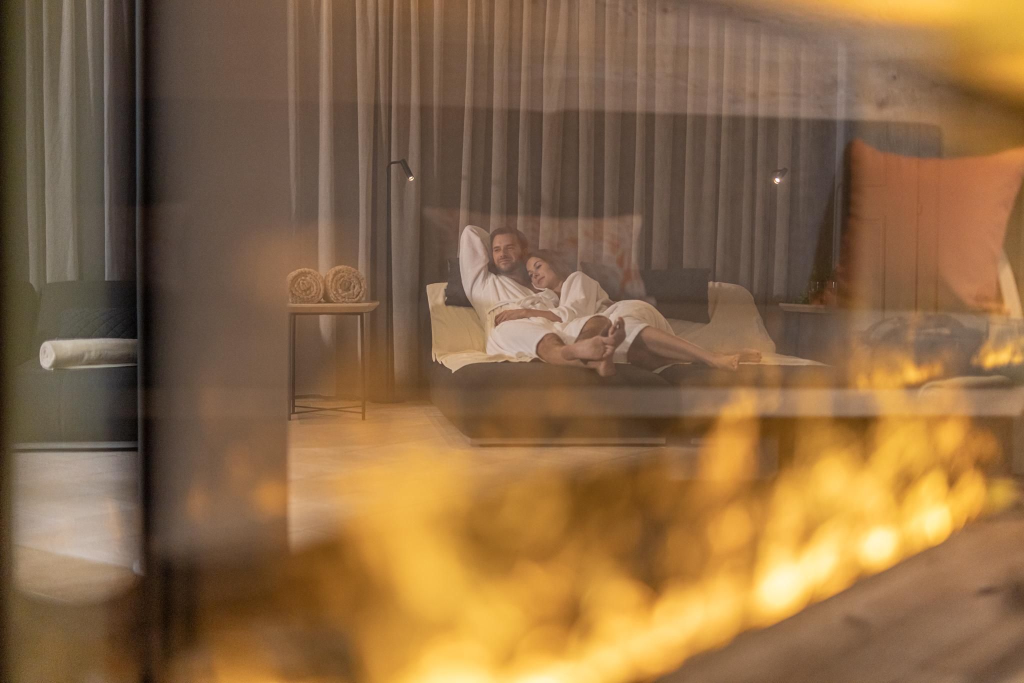 A cozy couple relaxes in a modern bedroom. In the foreground, an elegant fireplace creates a warm atmosphere.