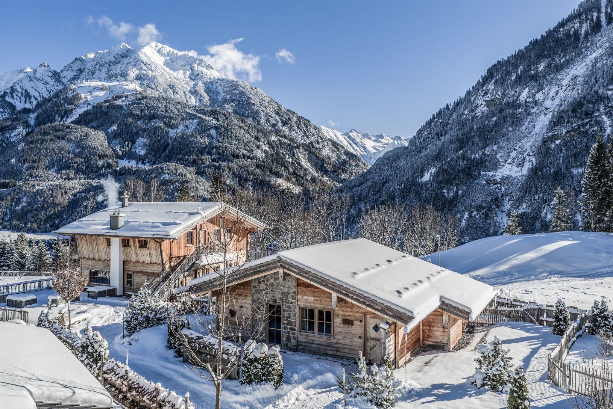 A winter landscape with snow-covered chalets and majestic mountains in the background. The sky is blue and the sun is shining on the snow-covered roofs.