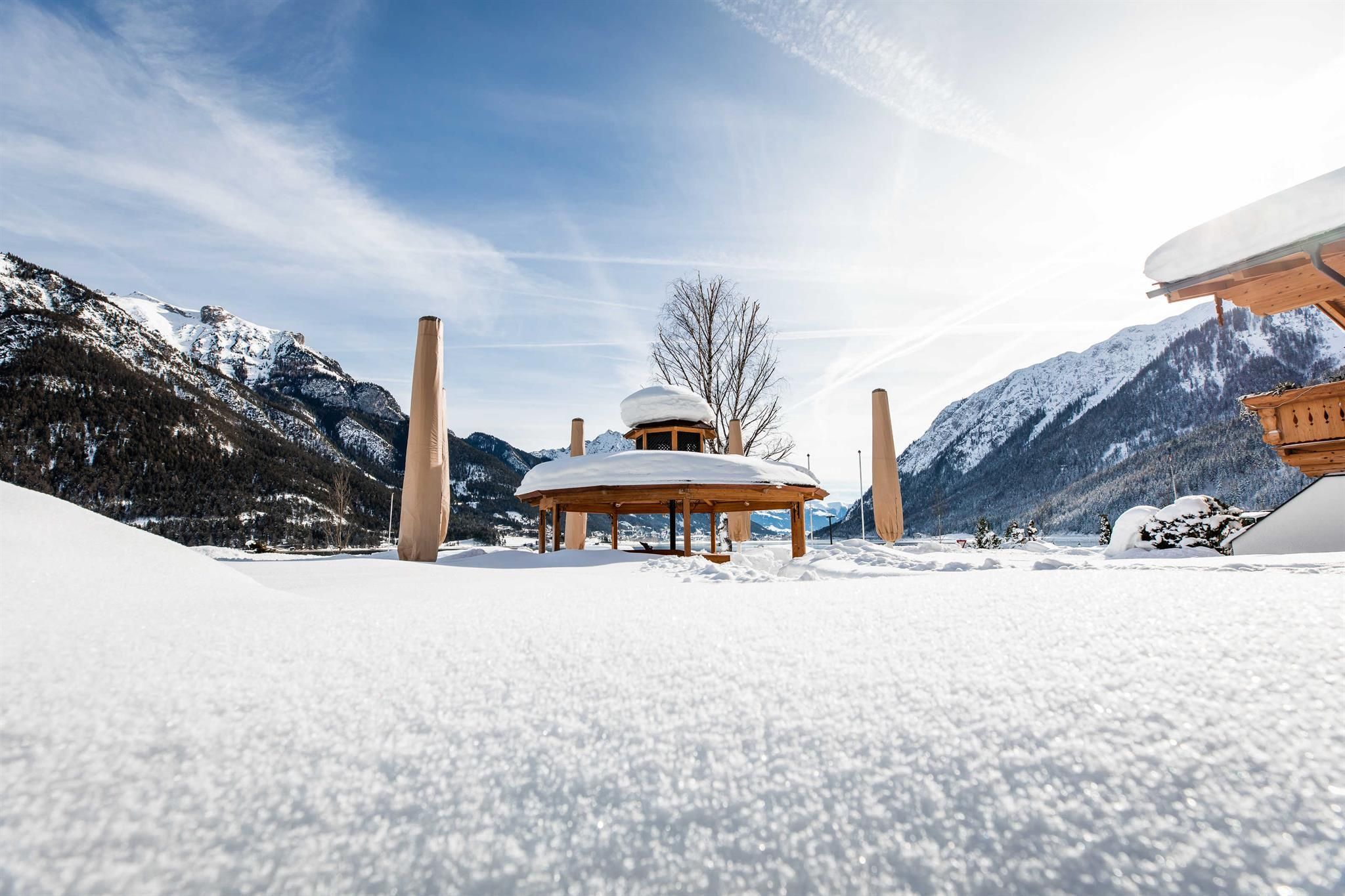 A snowy landscape with a modern wooden building and tall rocks in the background. The sky is clear and bright blue.