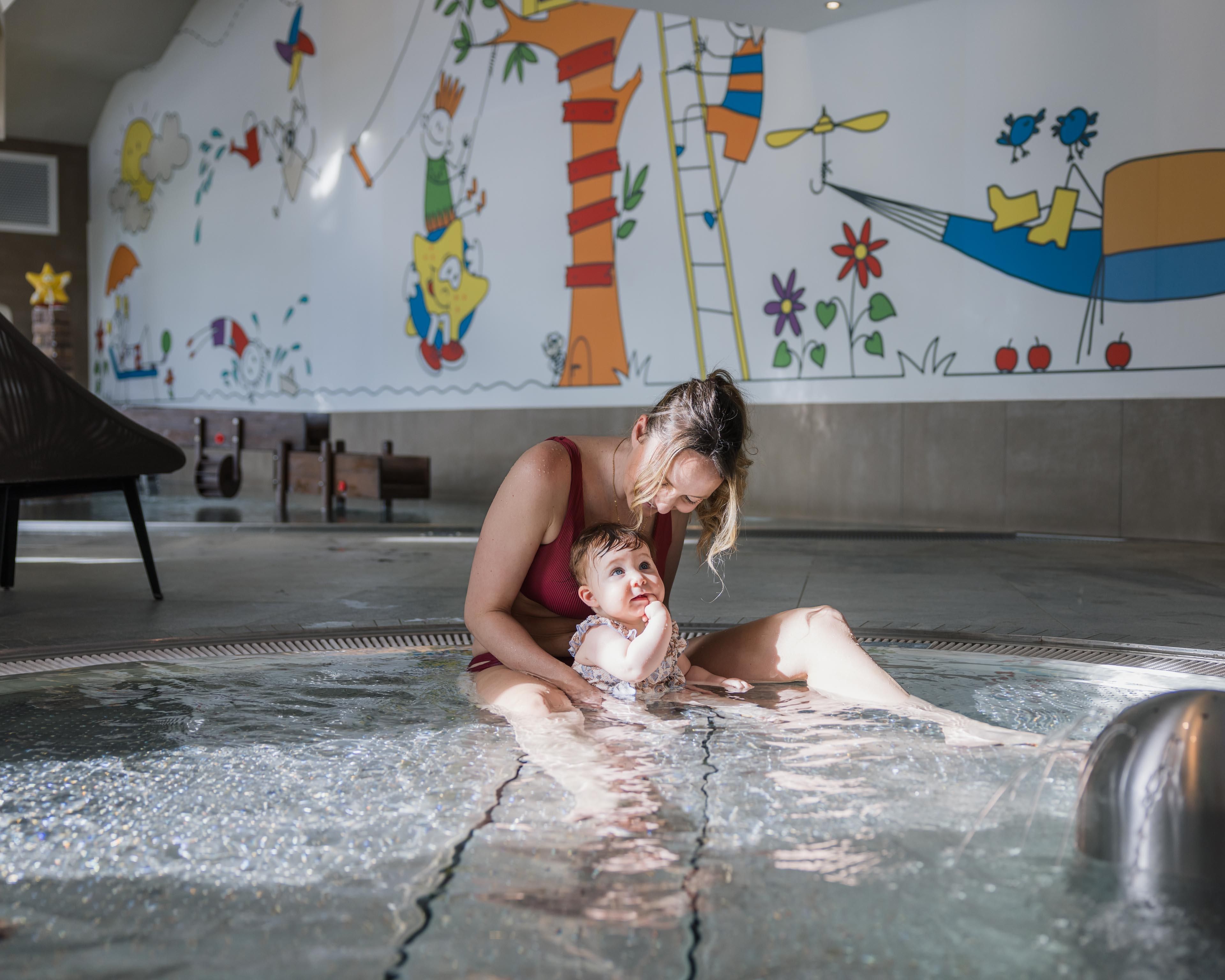 A mother plays with her baby in the water in the baby pool, where carefree splashing is guaranteed. In the background, a colorful mural can be seen.