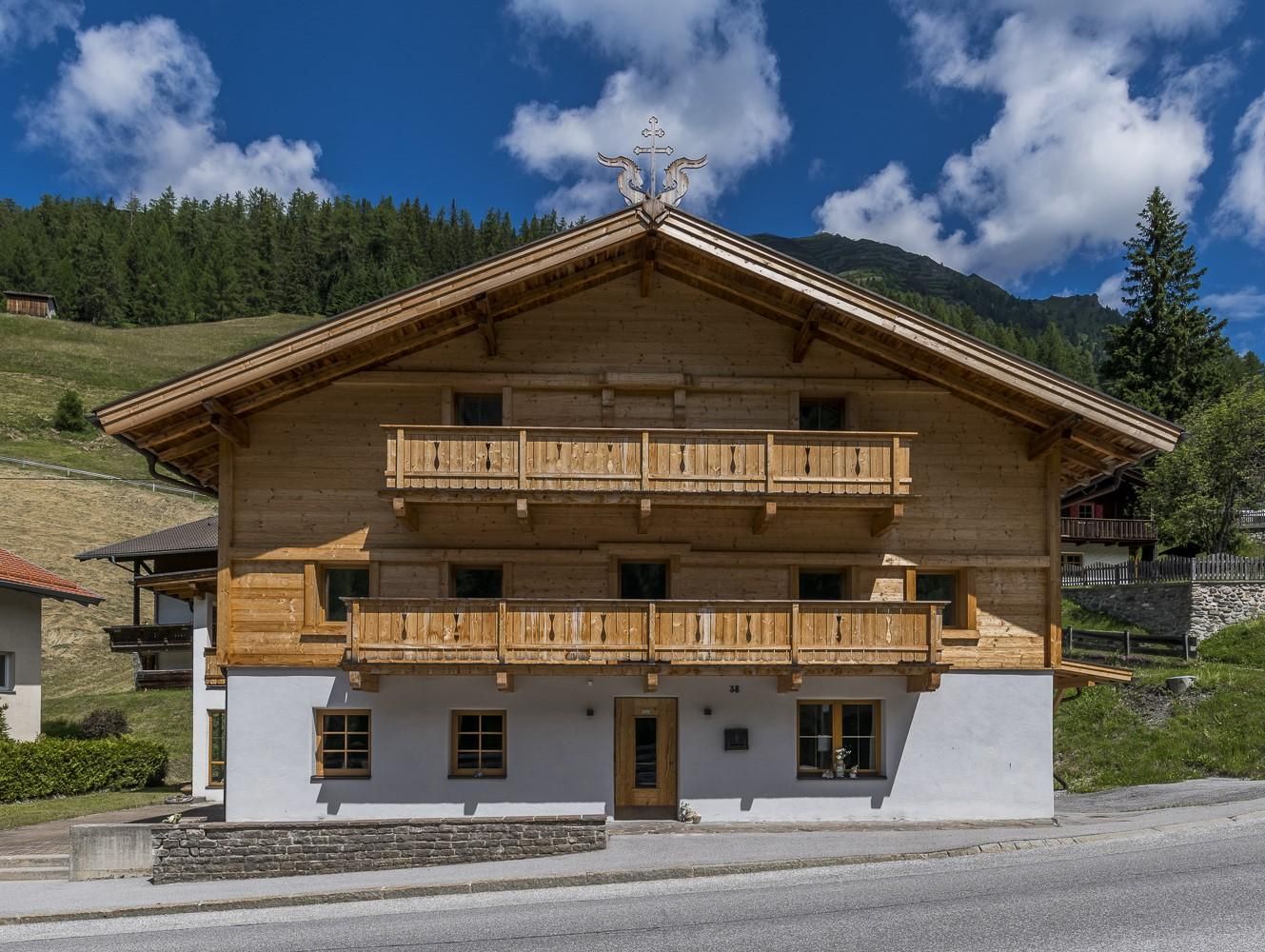 A traditional wooden house with a balcony in Alpine style. In the background, there are green hills and a blue sky with some clouds.