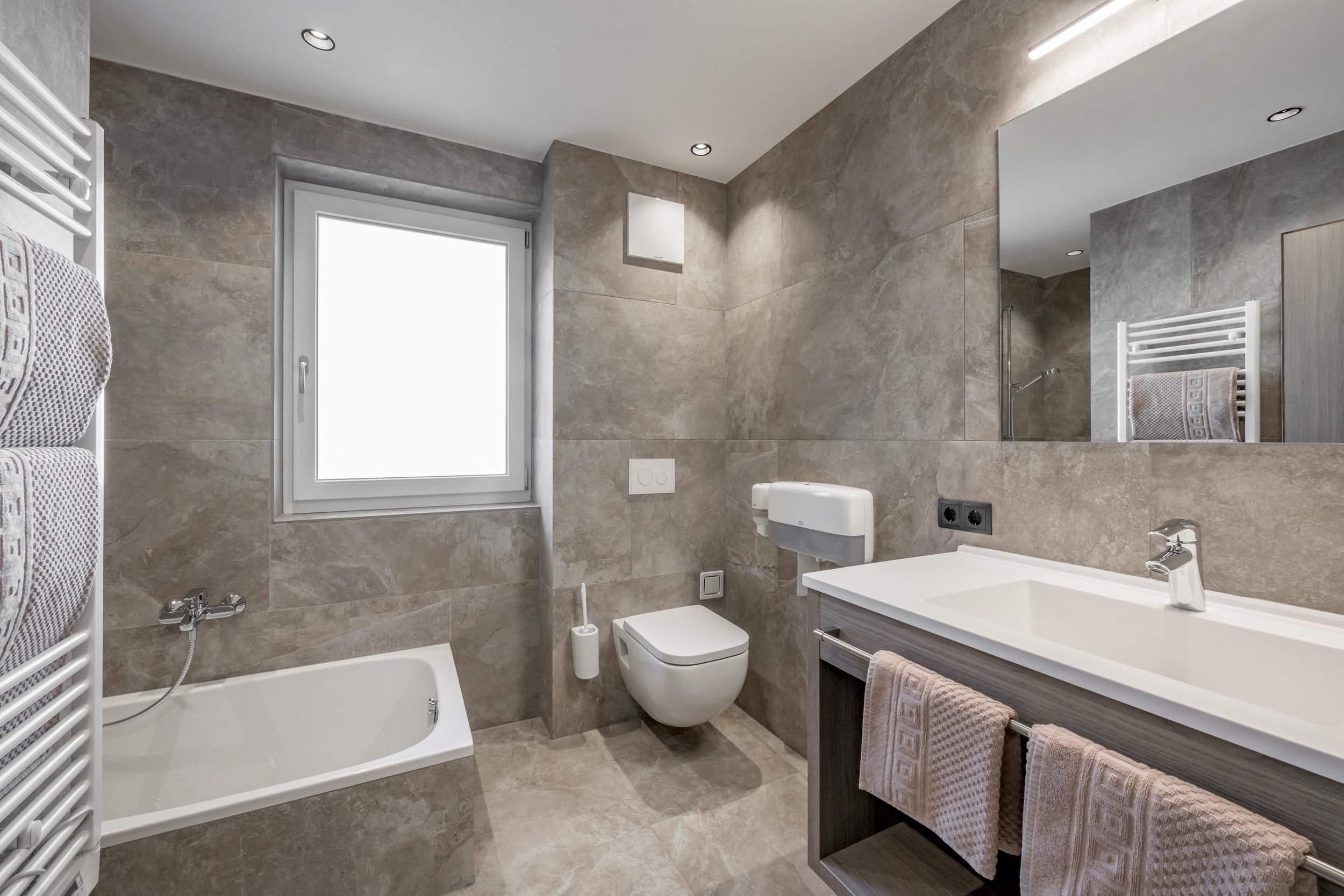 A modern bathroom with gray tiles and a bathtub. There is a wash area with a sink and towels.