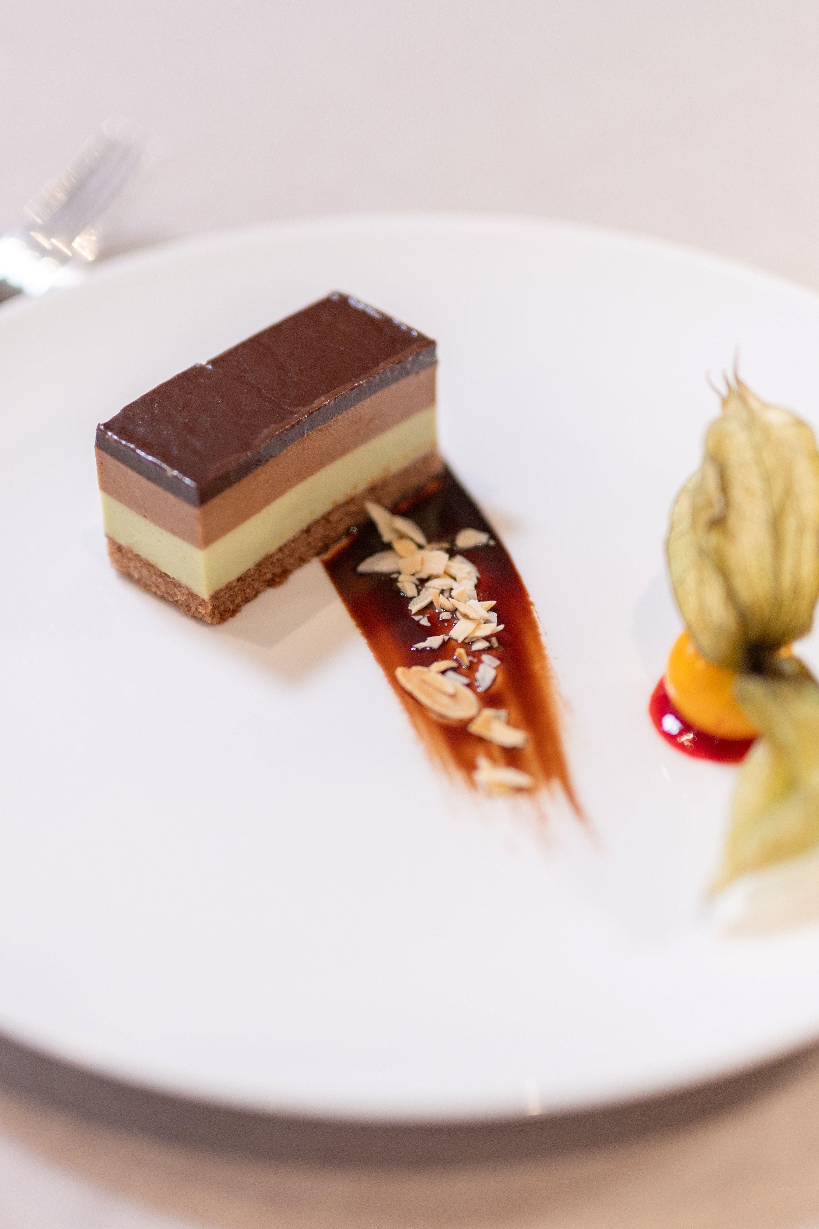 An elegant dessert on a white plate. It consists of several layers in different colors, adorned with a sauce and fruits.