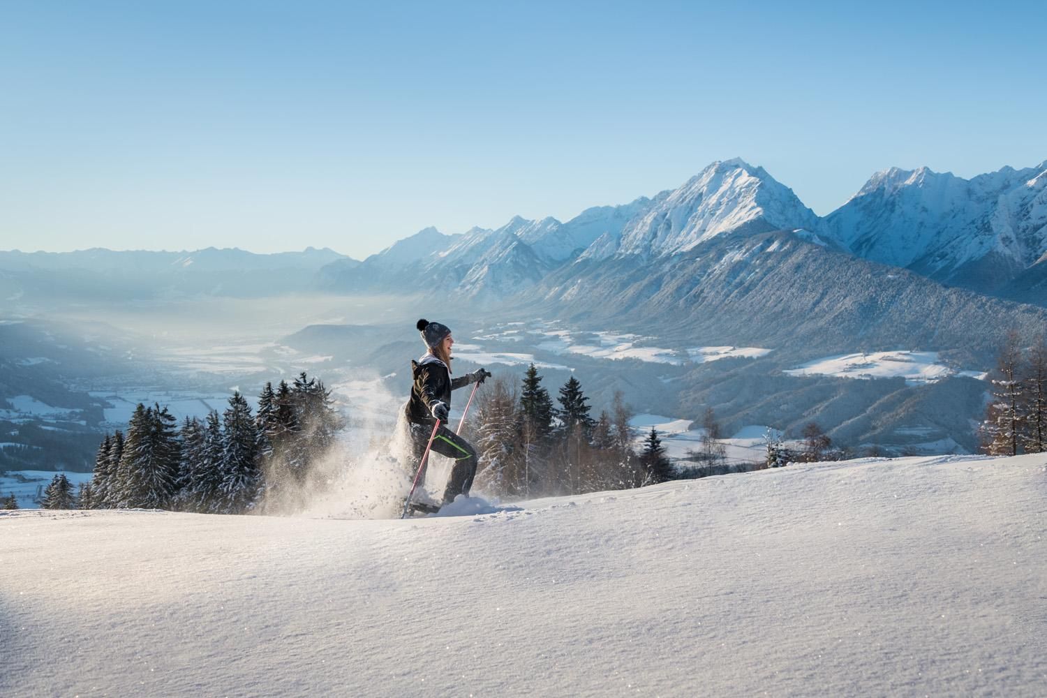 A person is walking in the snow with ski poles. In the background, impressive mountains and a clear sky can be seen.