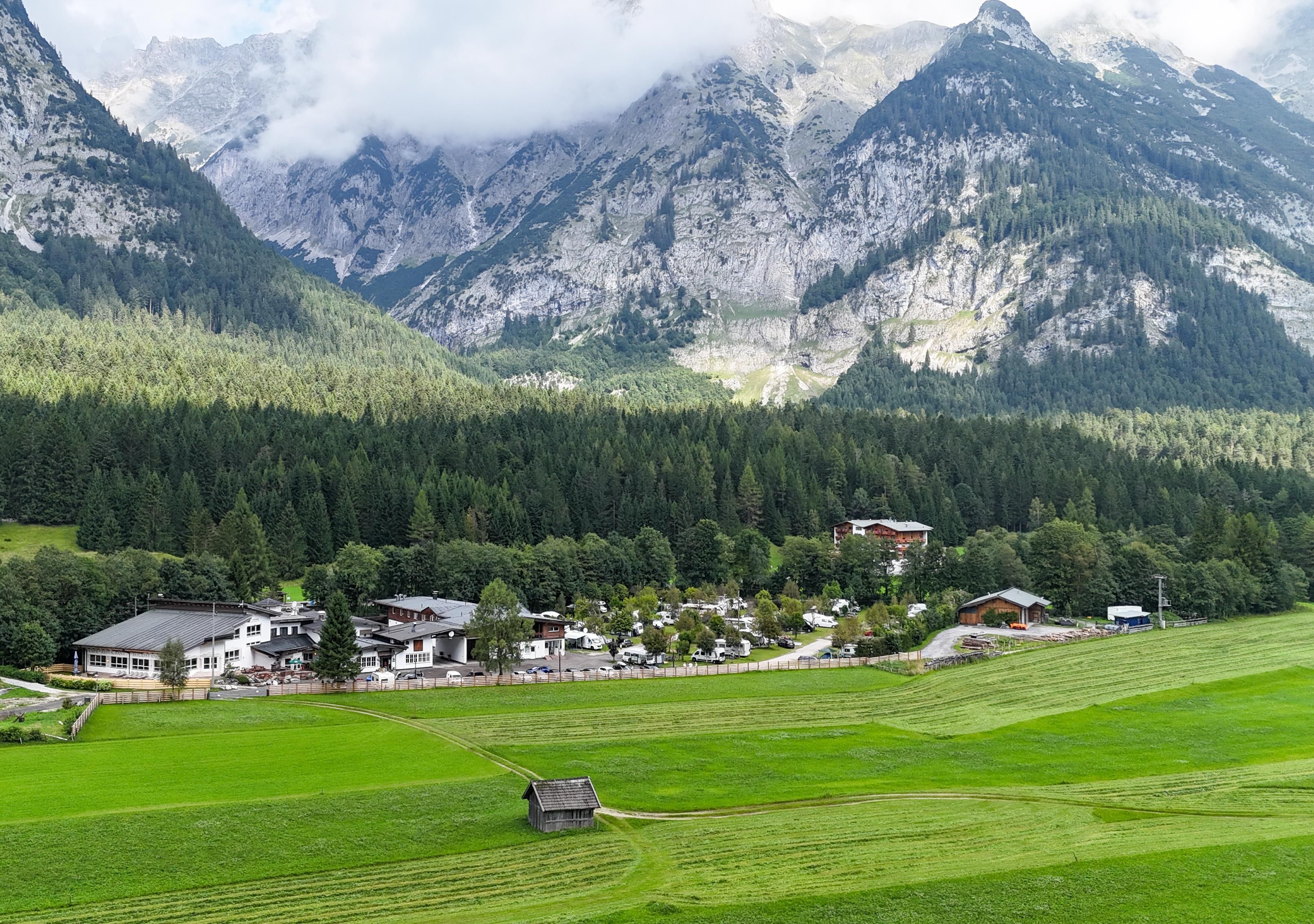 A picturesque landscape with green meadows and beautiful mountains in the background. In the foreground, some buildings and a small wooden shed can be seen.