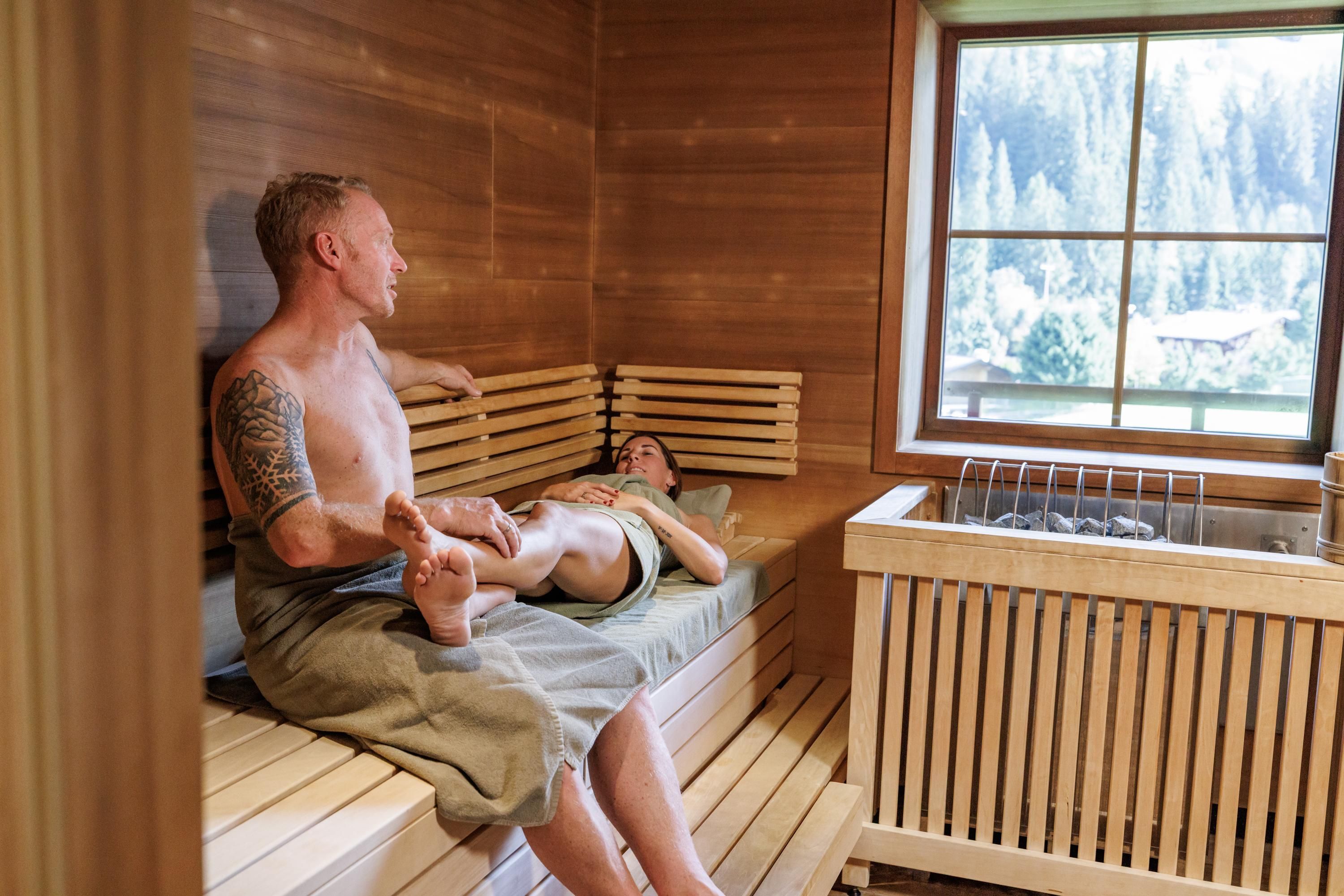 A cozy sauna room with wooden benches and a window view of nature. A couple relaxes and enjoys the tranquility.