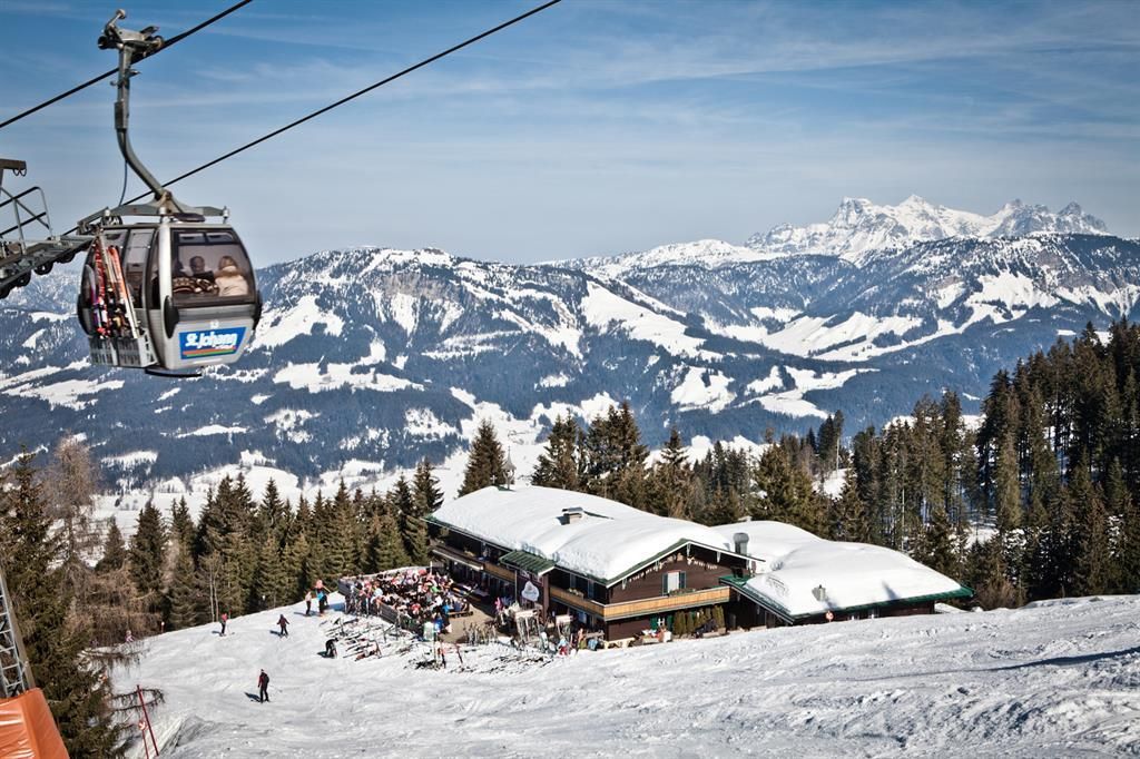 A mountain landscape with snow-covered hills and a ski lift. Below is a hut surrounded by skiers and snow-covered trees.