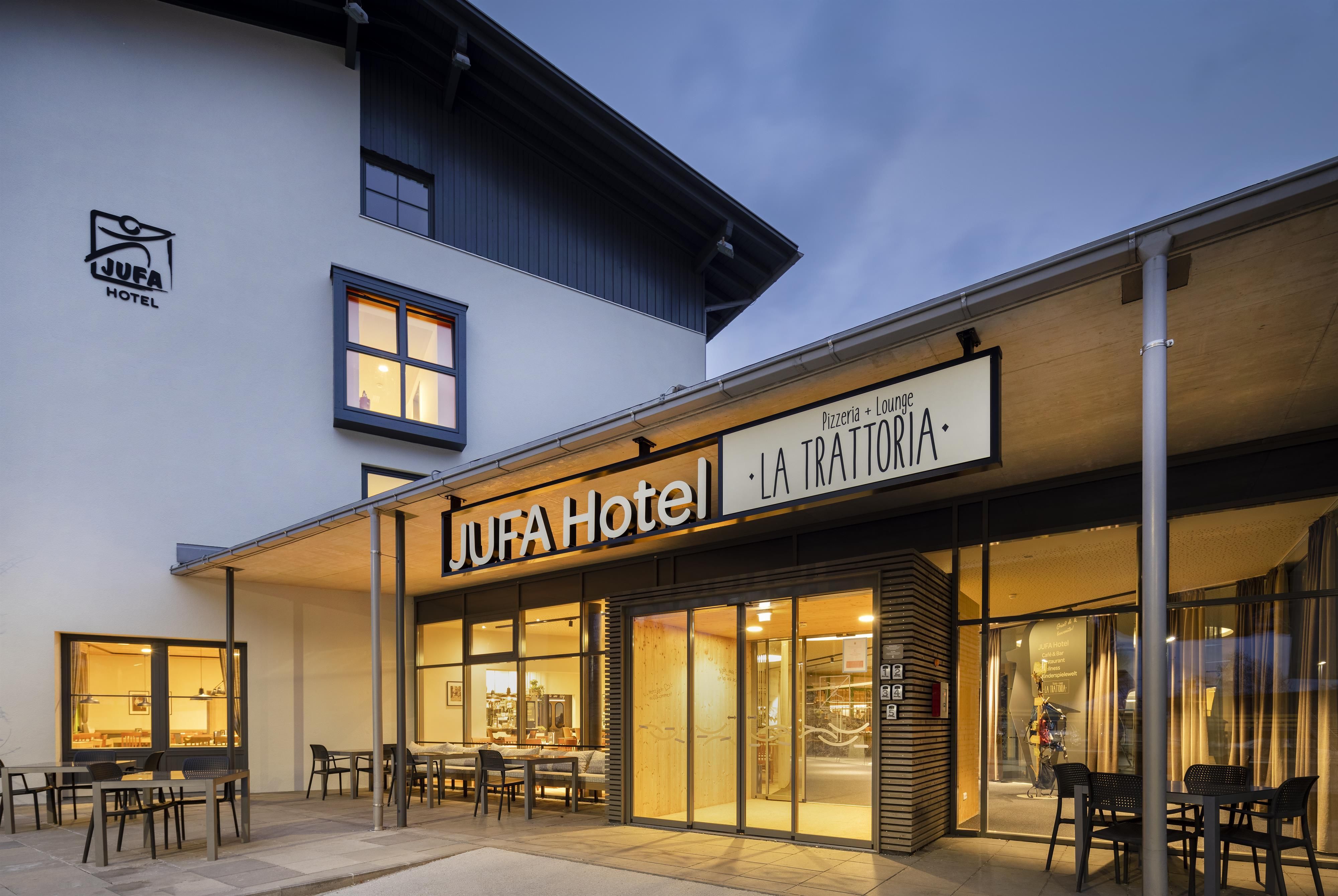 A modern hotel building with an inviting facade and large windows. Outside, there are tables and chairs for guests.